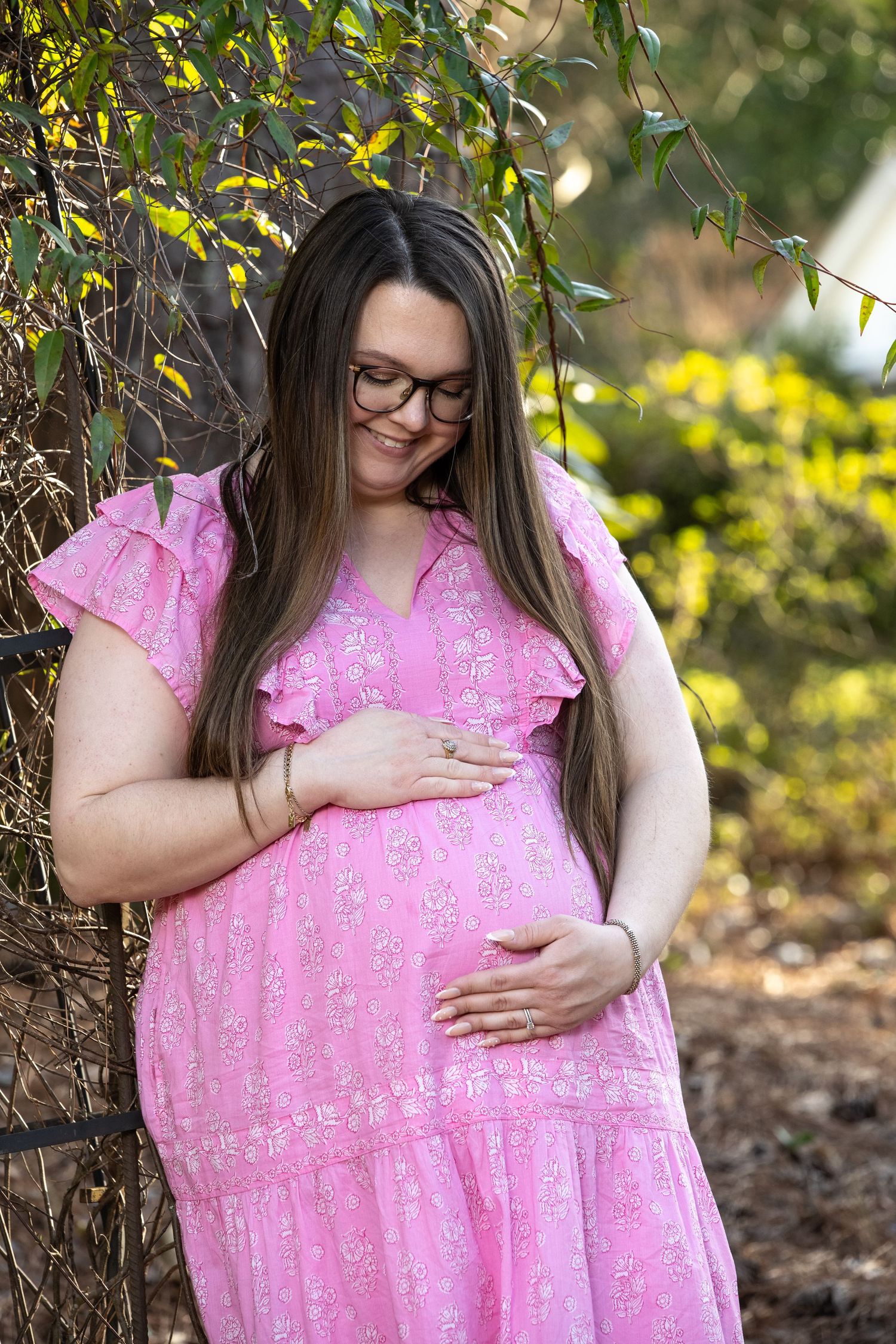 Expecting mother in pink floral dress and glasses gently holds her baby bump while standing near trees in a garden.