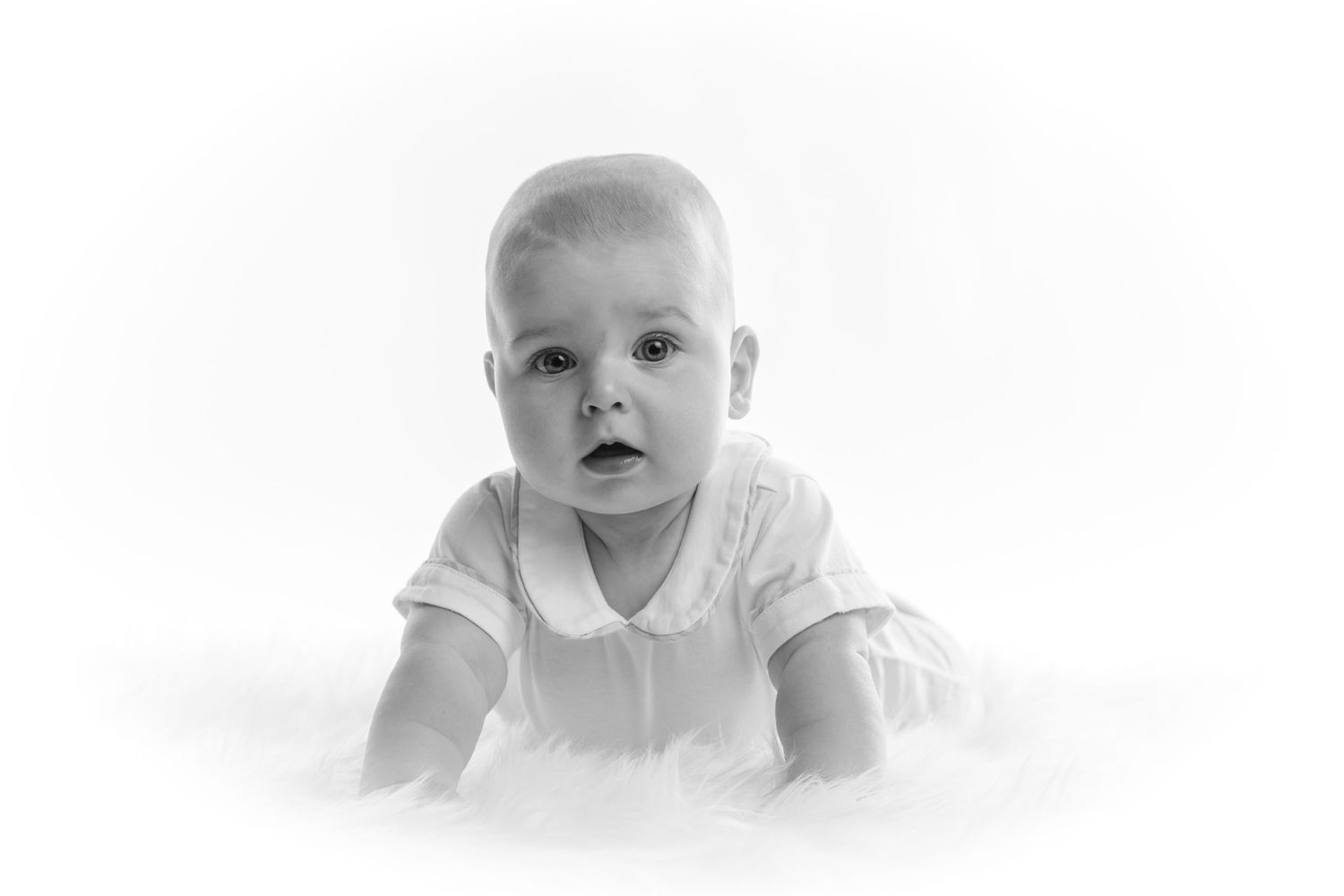 Black and white portrait of an adorable baby in white outfit doing tummy time on a soft white surface.