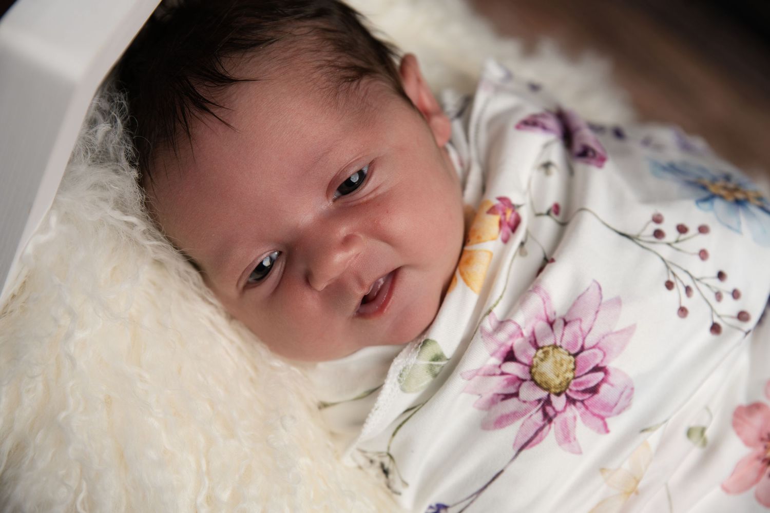 Newborn baby swaddled in a floral blanket lying on a fluffy white surface, with dark hair and alert eyes.