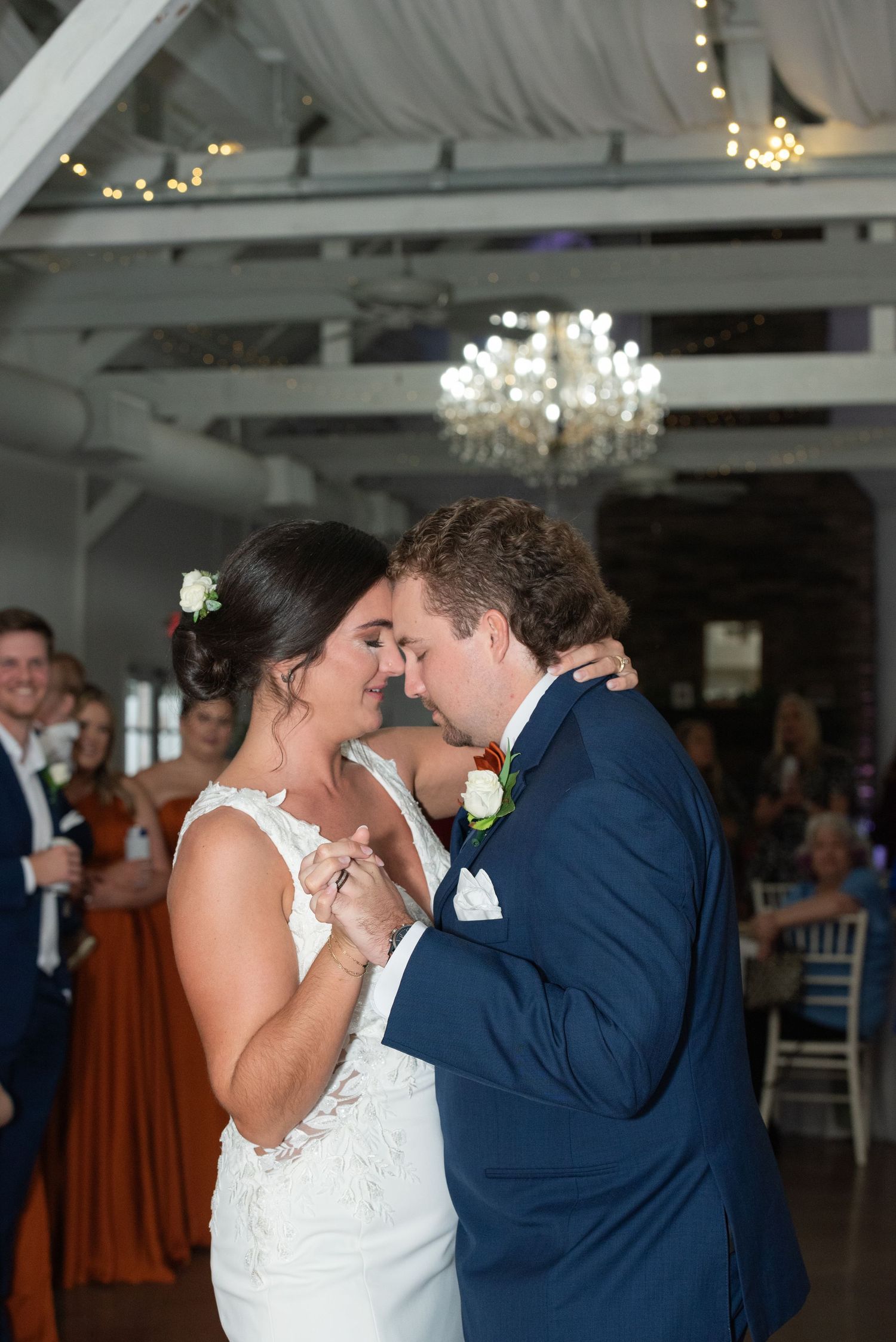 Bride and groom share their first dance in an elegant white venue with chandelier lighting and guests watching.