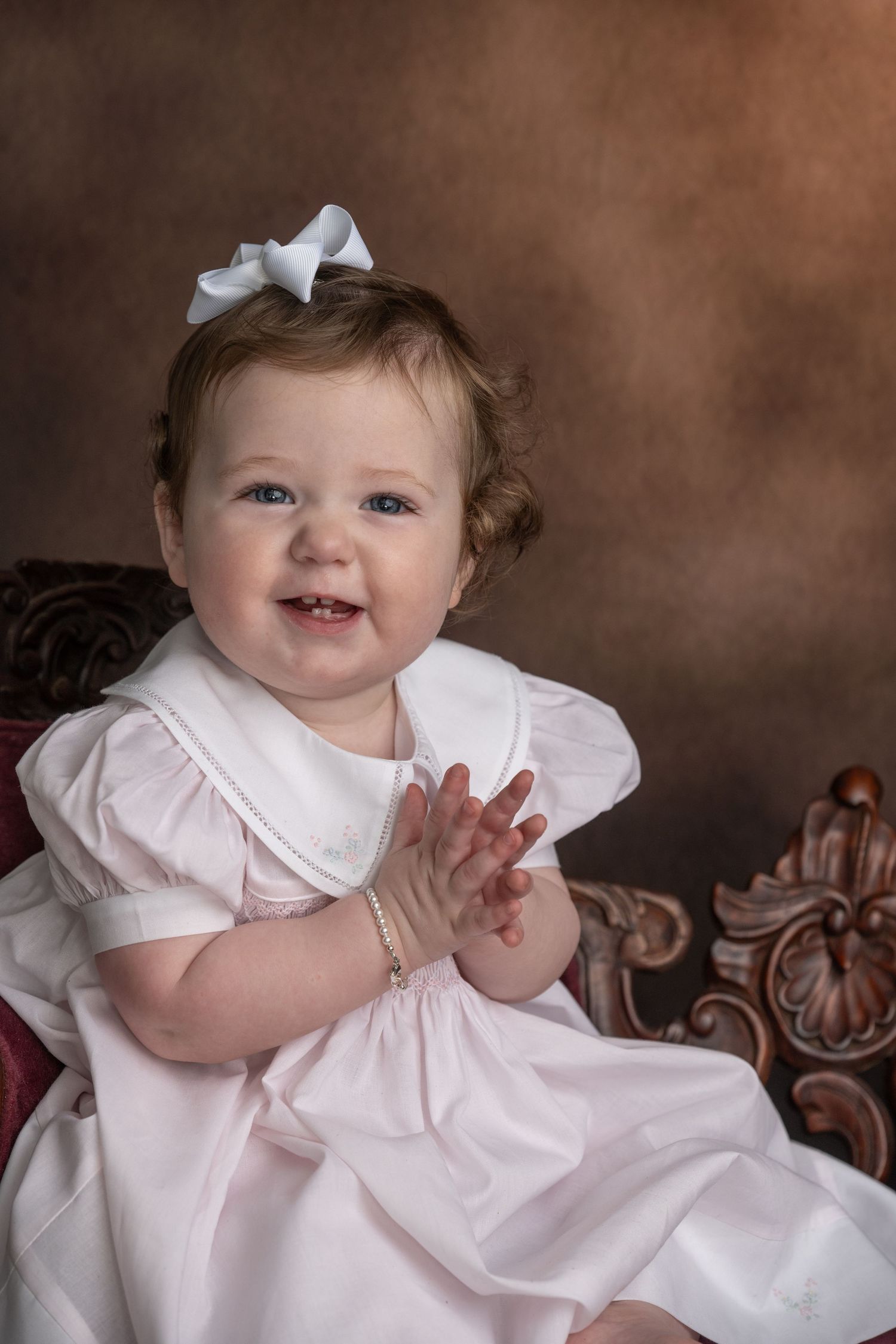 Smiling baby girl in pink dress with white bow clapping hands, seated in ornate chair for professional portrait.