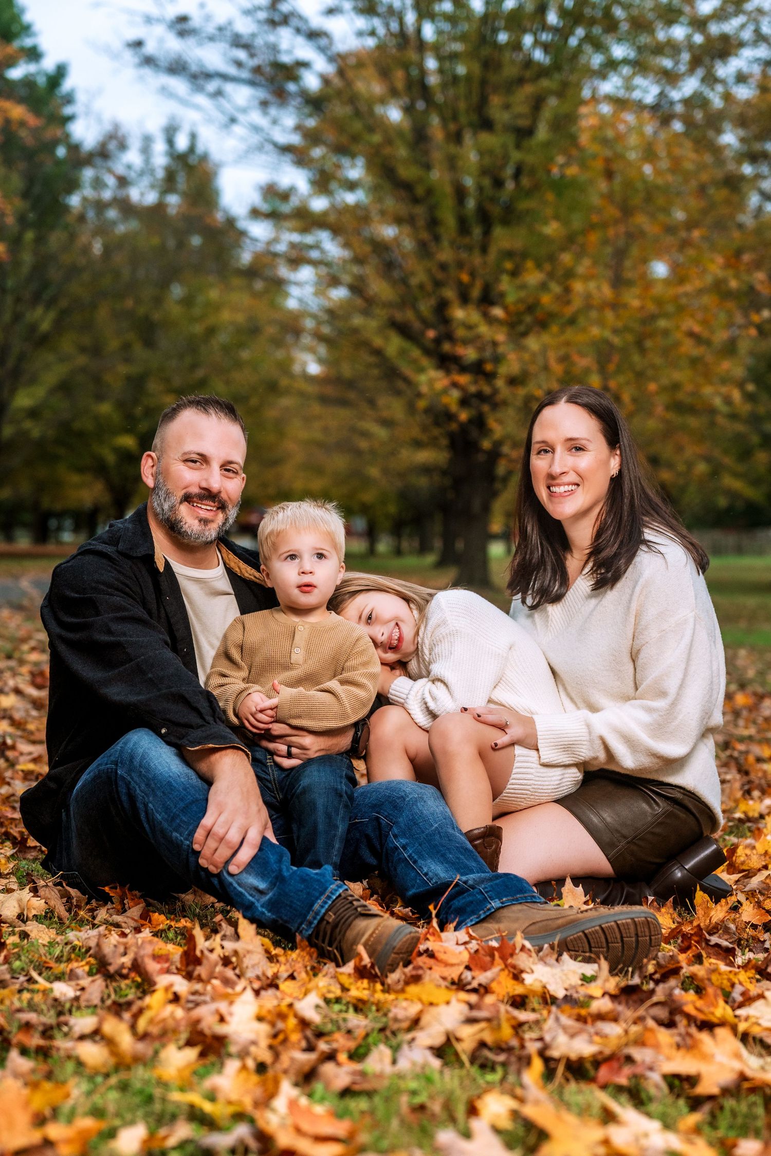 Family of four sitting on autumn leaves in a park, smiling together surrounded by colorful fall foliage.