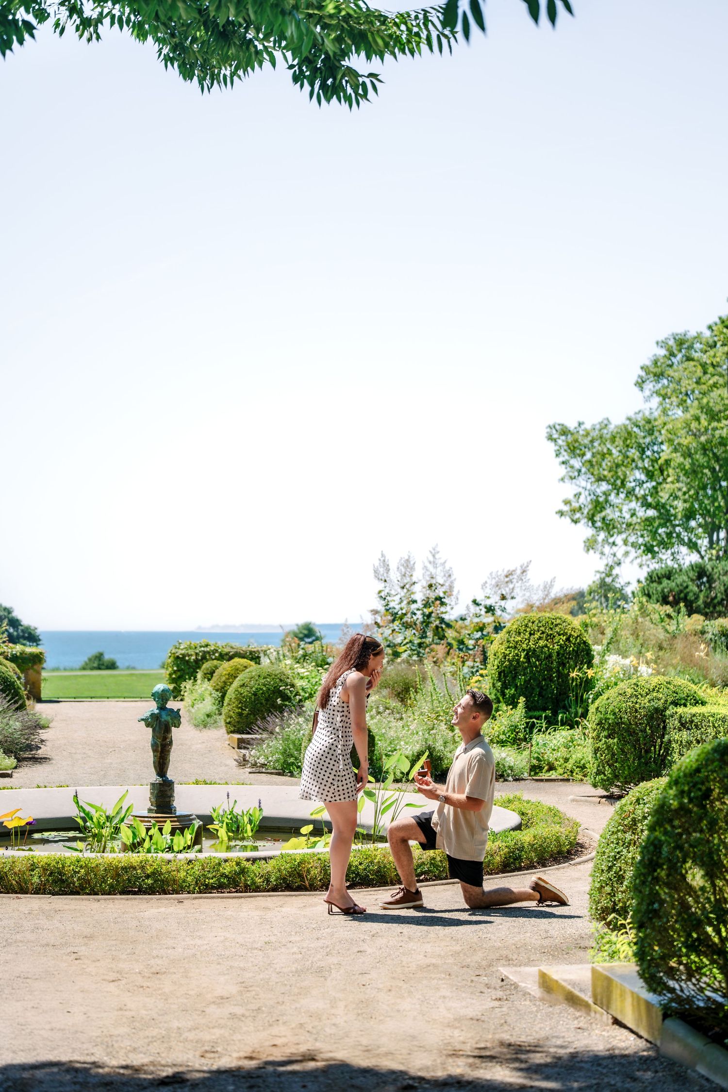 Man proposes to woman on one knee in a beautifully manicured garden with ocean views on a sunny day.