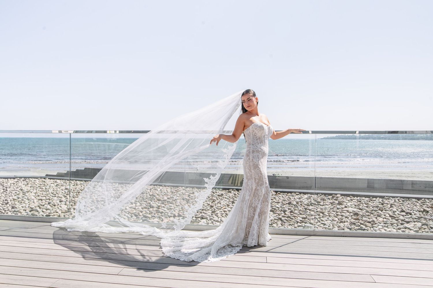Bride in fitted lace gown with flowing veil blowing in wind on seaside deck under clear blue sky.