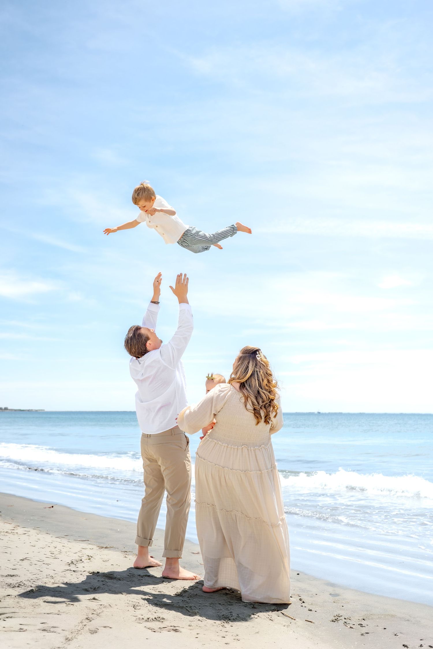 Family enjoying beach day as father tosses young child in the air while mother watches, all dressed in white and beige.