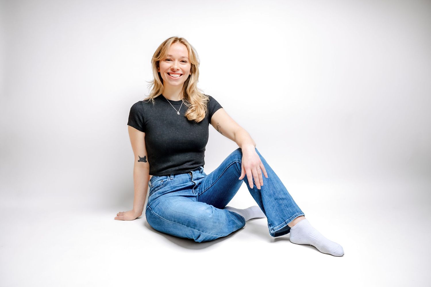 Young blonde woman smiling, seated on floor in black t-shirt, blue jeans, and white socks against white background.
