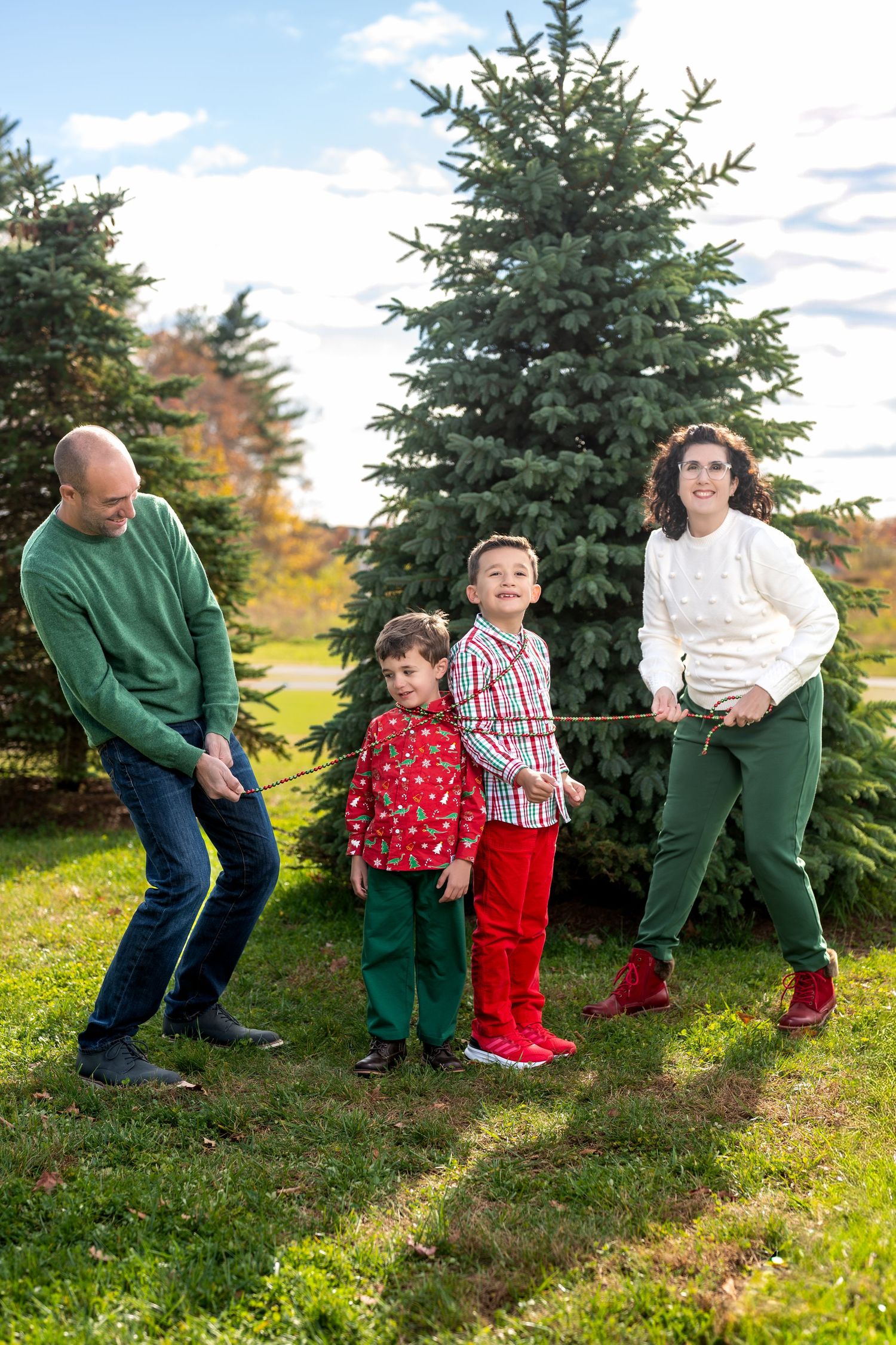 Family of four playing tug-of-war outdoors near a large evergreen tree, dressed in festive holiday attire.