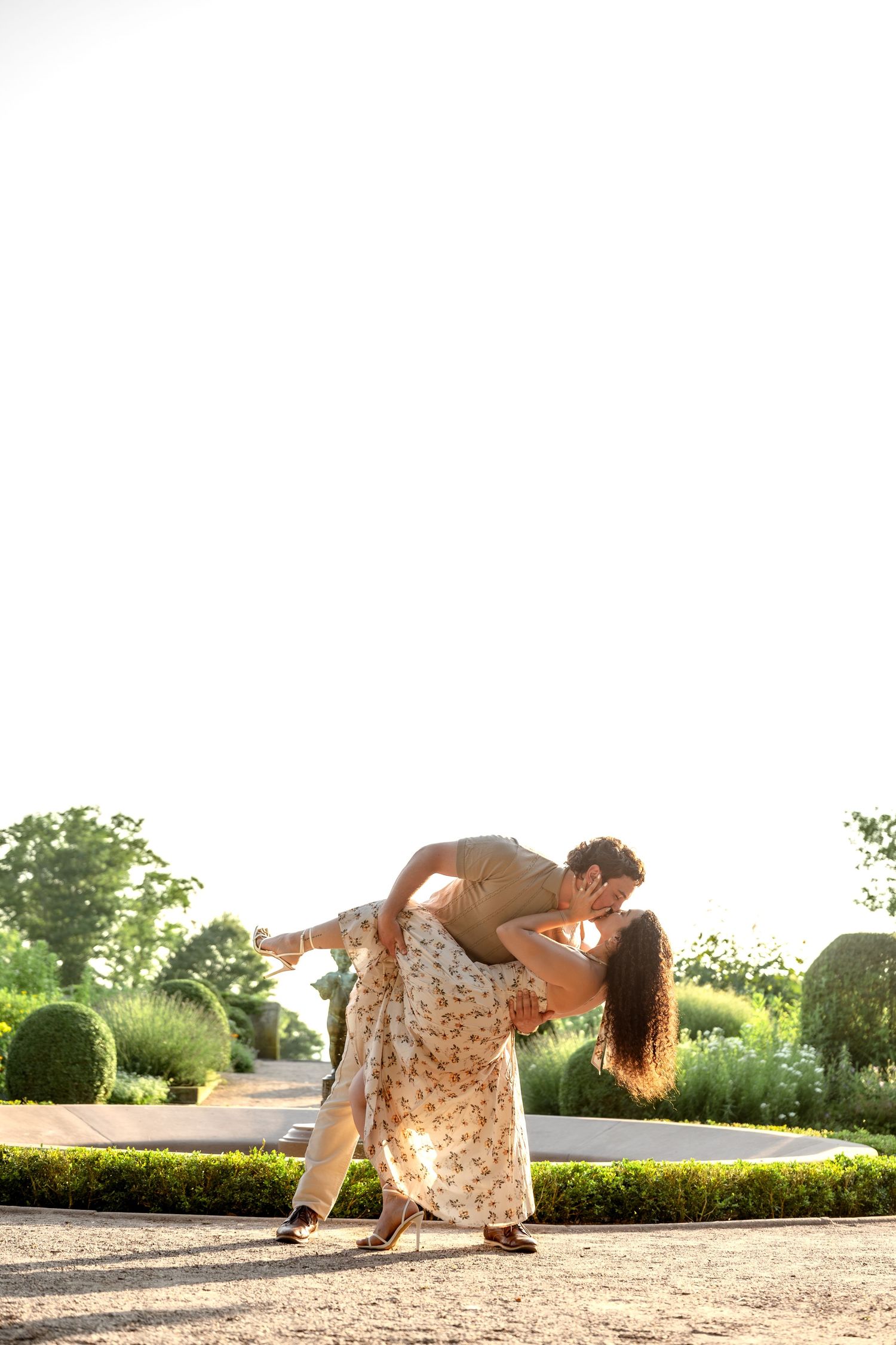 Couple sharing a romantic dip kiss in a sunlit garden with manicured hedges and greenery.