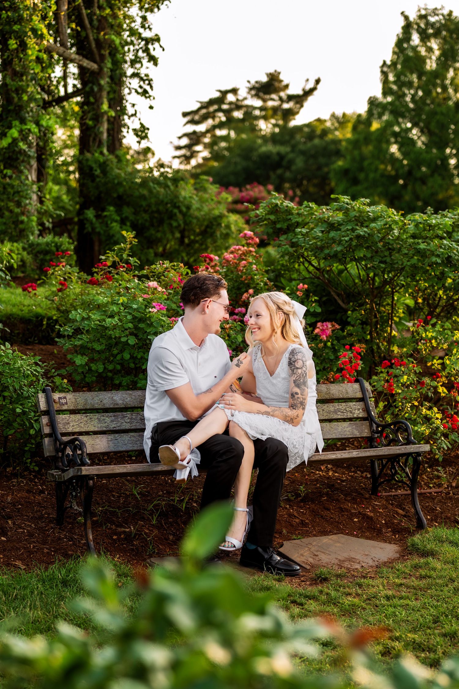 Couple sitting together on a wooden bench surrounded by blooming roses and greenery in a garden.