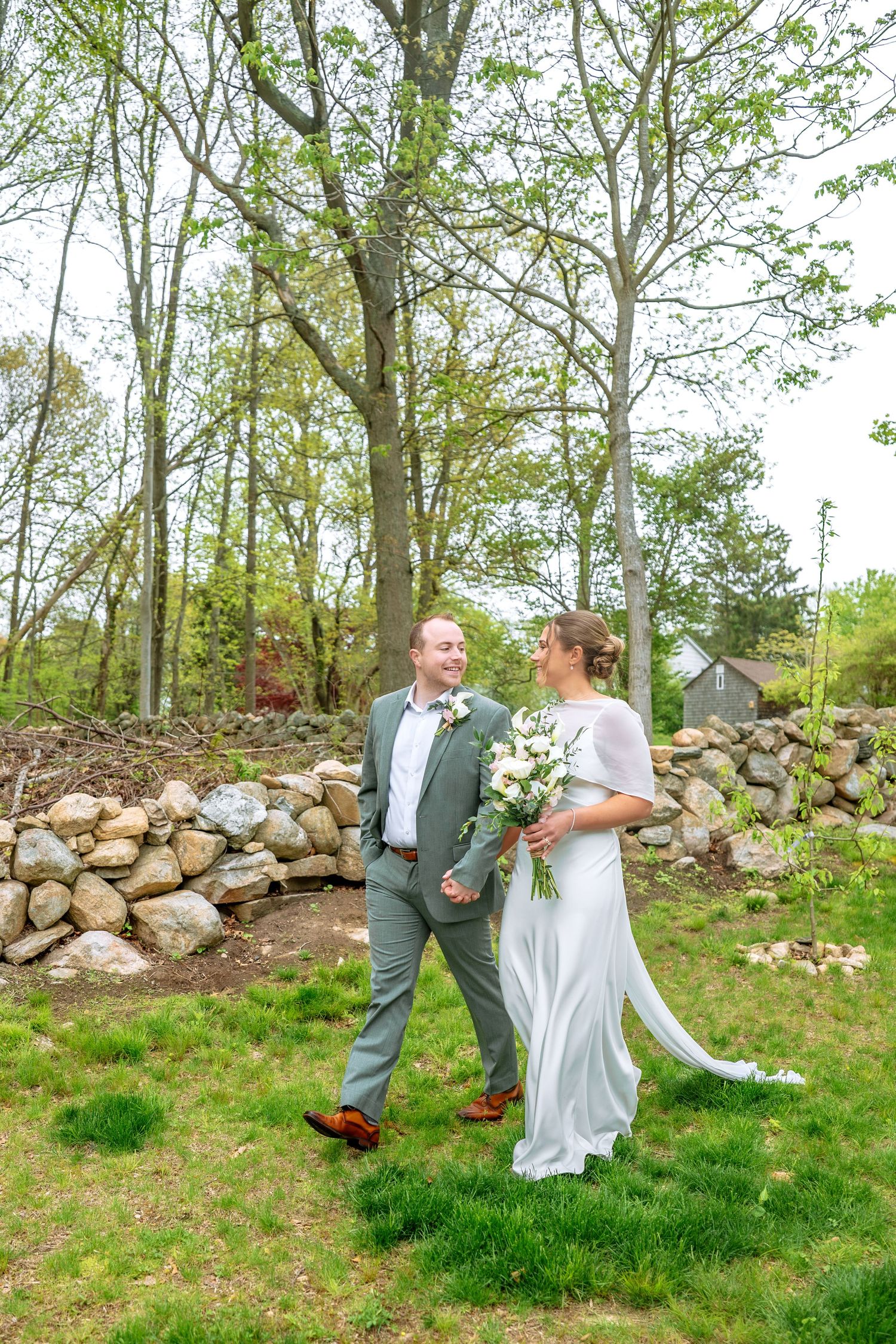 Bride and groom walk hand in hand on green grass, surrounded by stone walls and trees, as bride holds white bouquet.