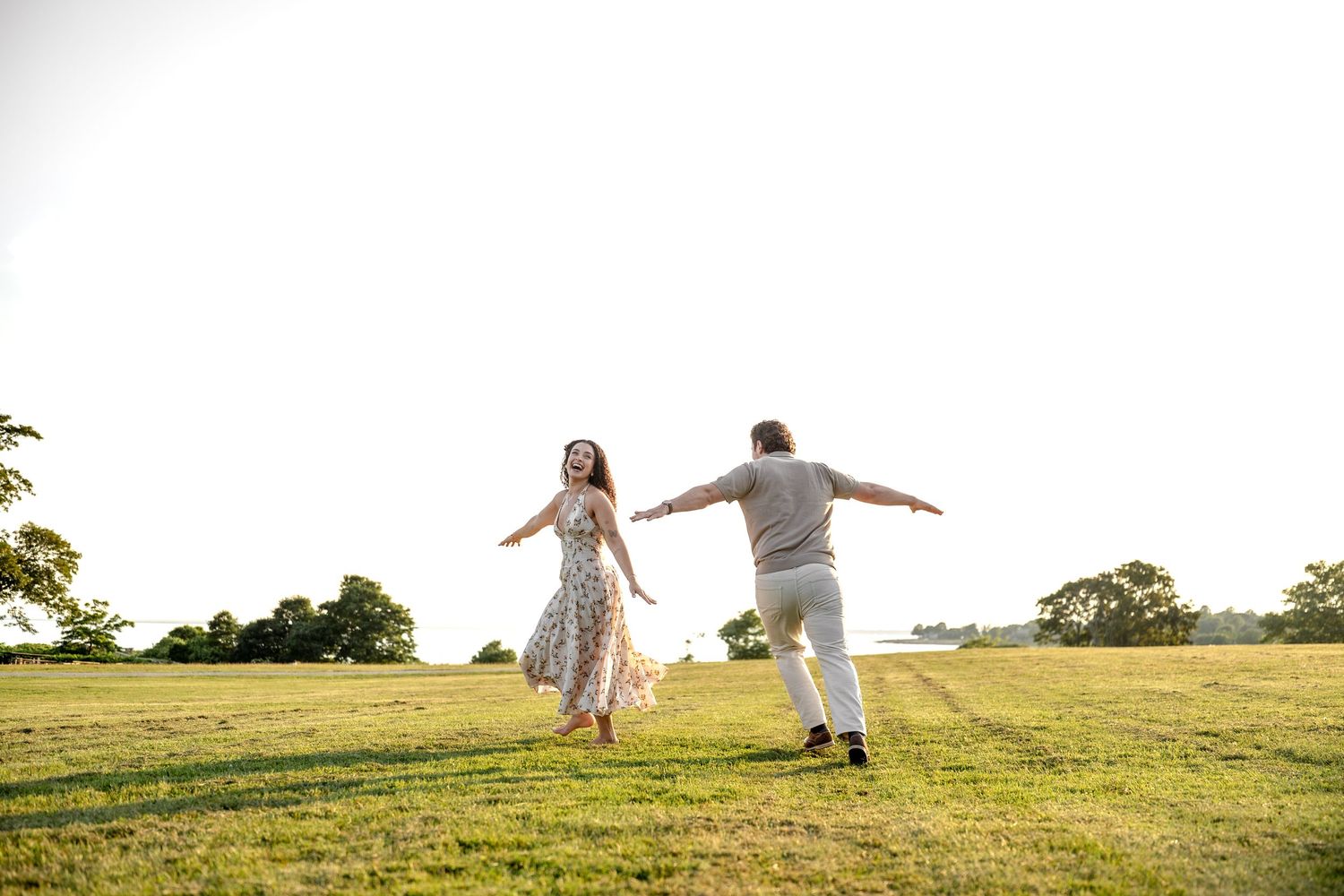 Couple dancing joyfully in a sunlit green field, woman in floral dress, man in white, trees in background.