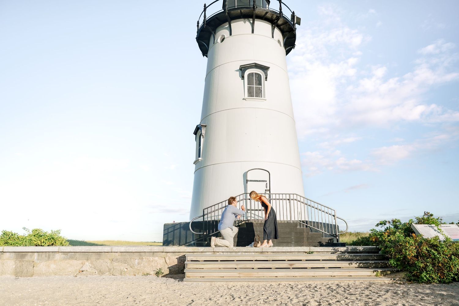 Couple embracing at the base of a white lighthouse on a sandy beach under a clear blue sky.