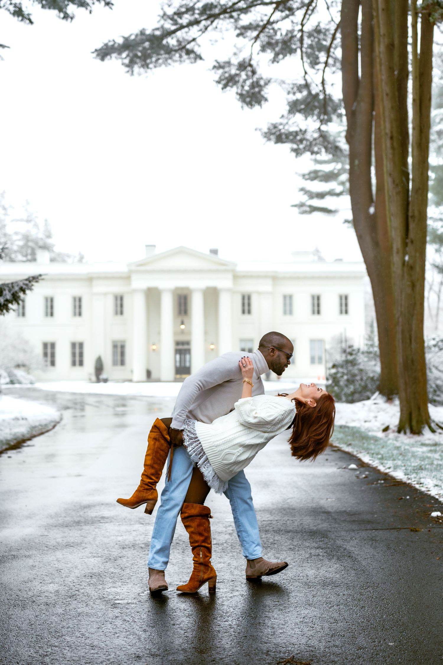 Couple dancing in the snow outside a white mansion, woman in brown boots dips back gracefully.