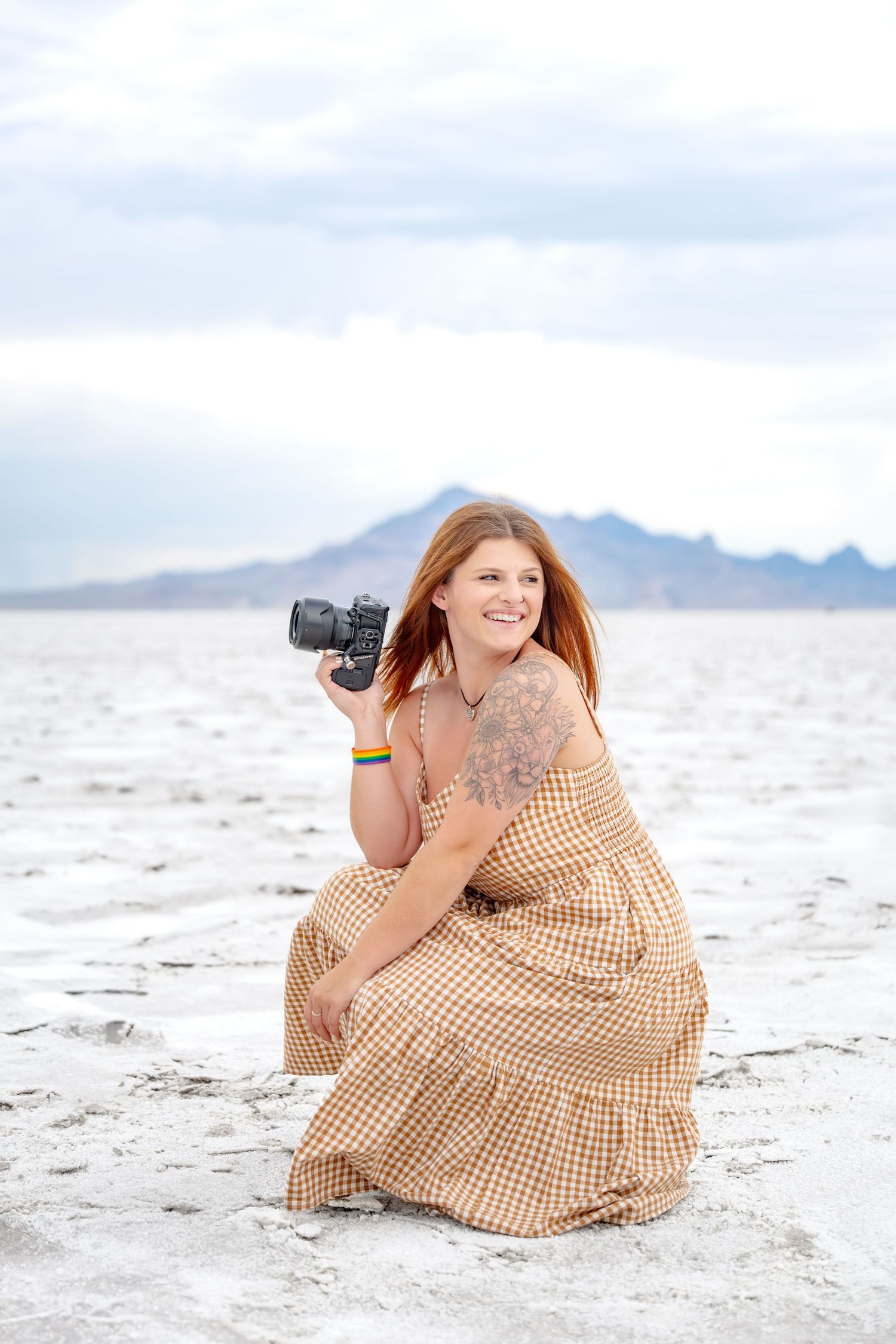 Woman in plaid dress crouching on salt flats holding camera, mountains in background.