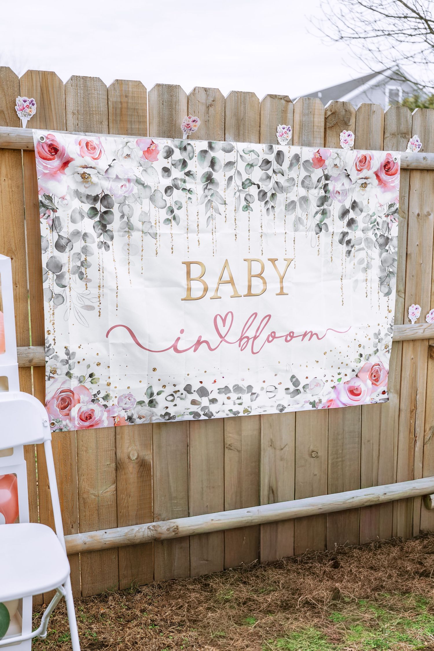 Floral baby shower backdrop banner reading Baby in Bloom with pink roses and greenery hanging on a wooden fence.