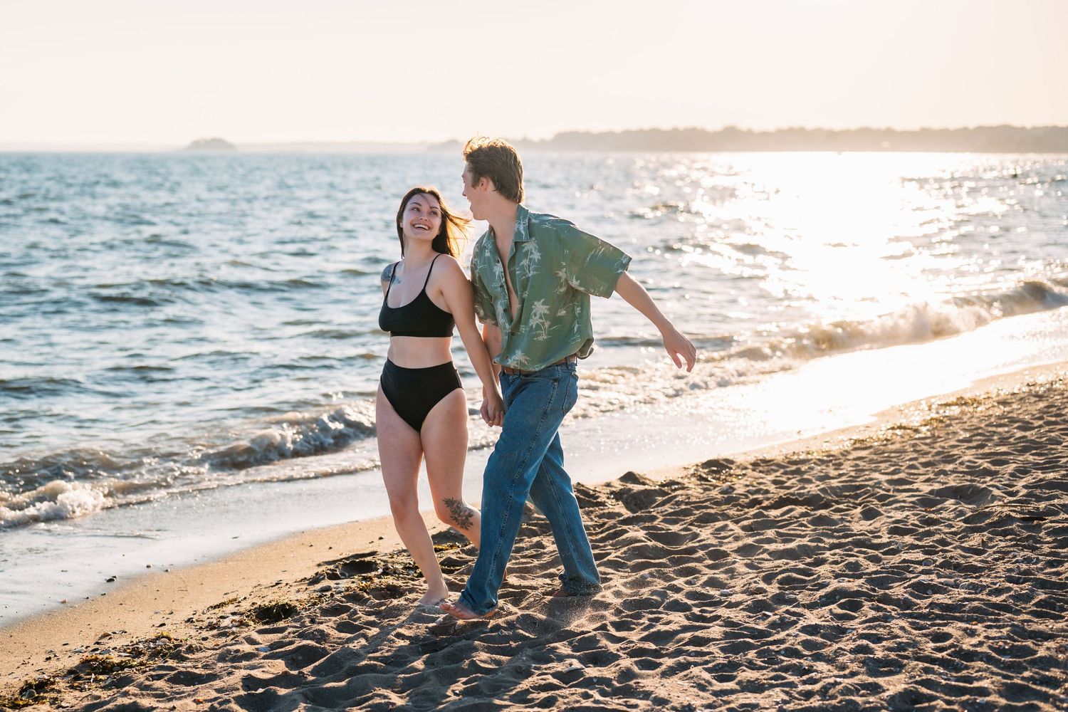 Couple walking along sandy beach at sunset holding hands.