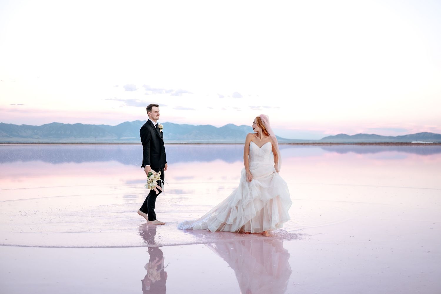Bride and groom walk on reflective pink salt flats at sunset, mountains in the background.