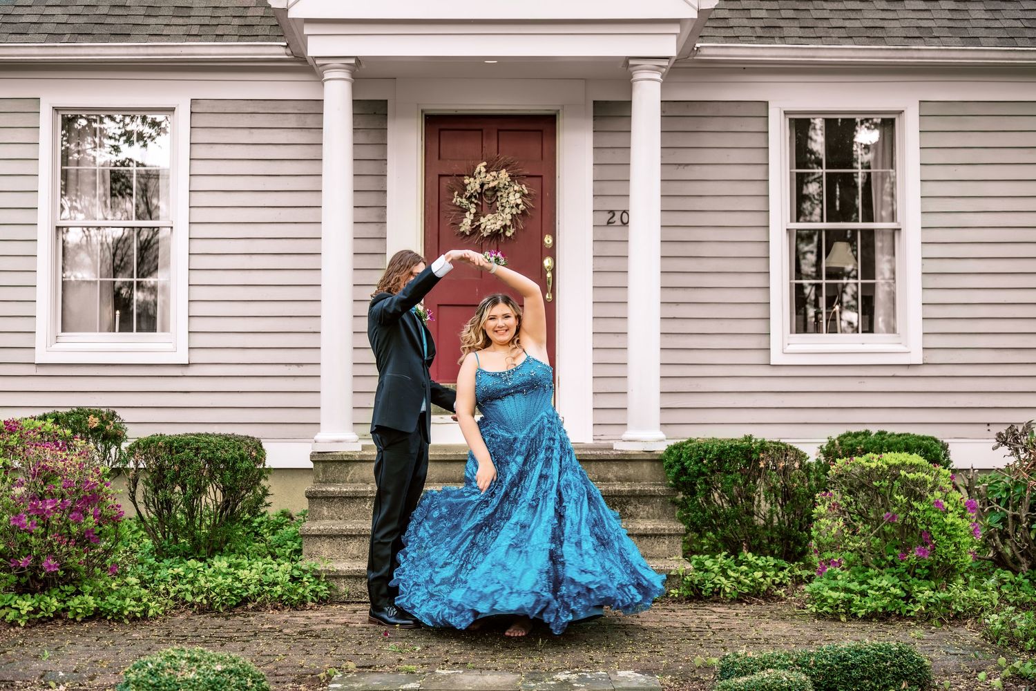 Couple dancing in formal wear outside a charming house, woman in blue gown twirling elegantly.