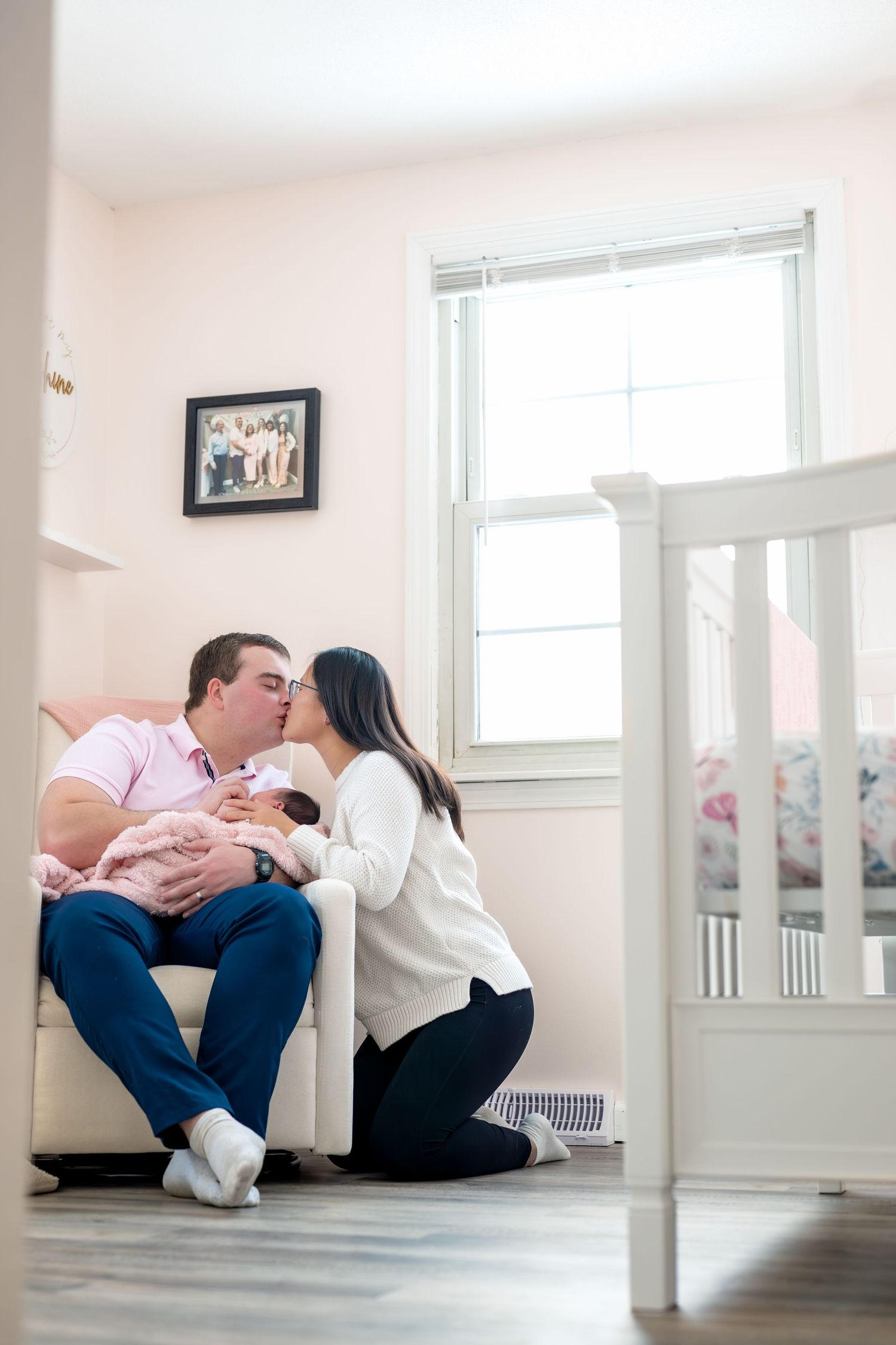 A couple shares a kiss while holding their newborn baby in a nursery room with a white crib nearby.