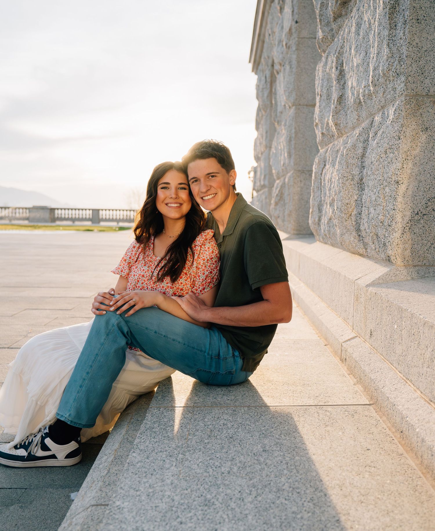 A young couple sits closely together on stone steps, smiling warmly, with a sunlit waterfront backdrop.