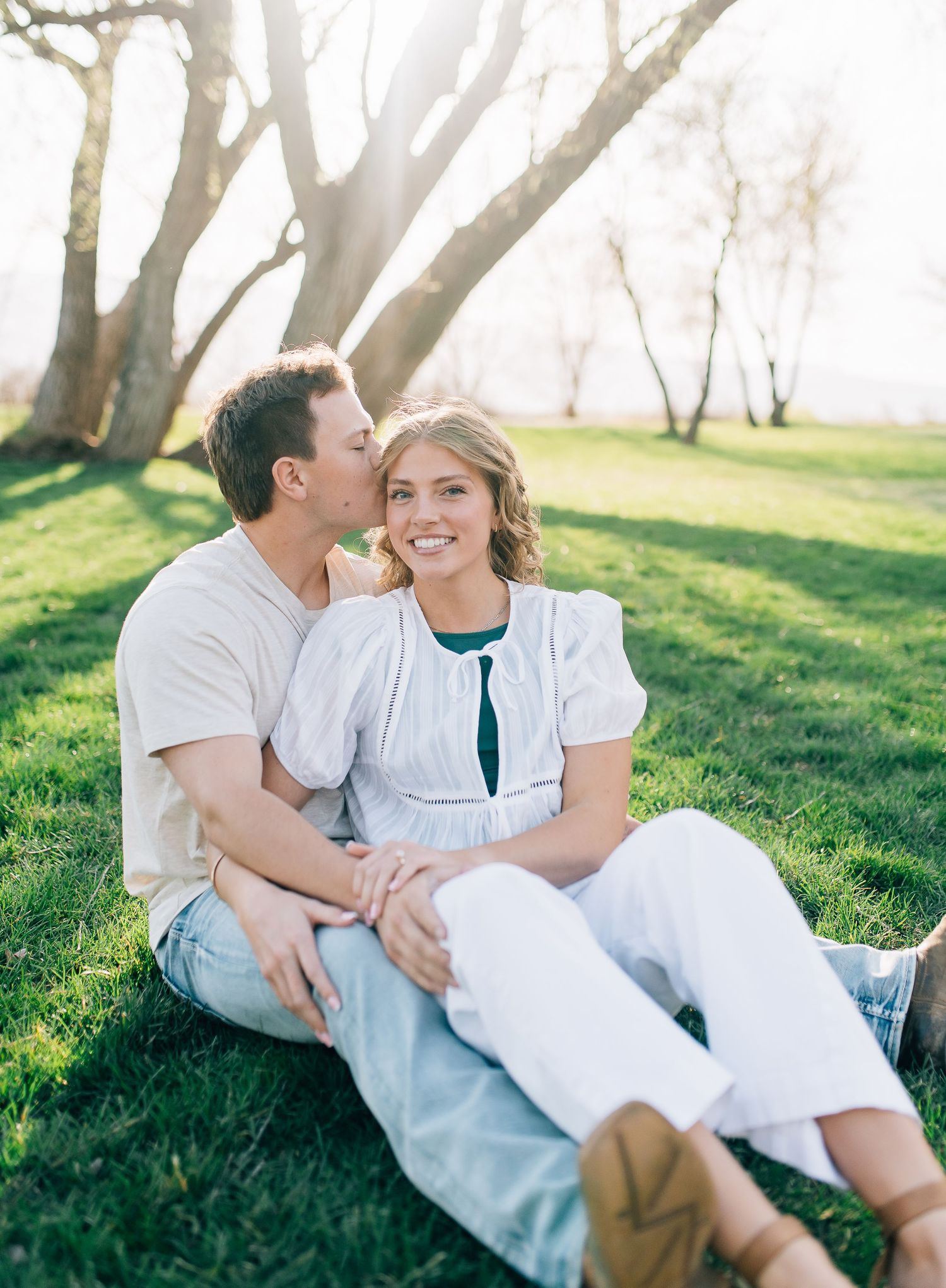 A loving couple relaxes together on green grass under trees in a sunlit park during springtime.