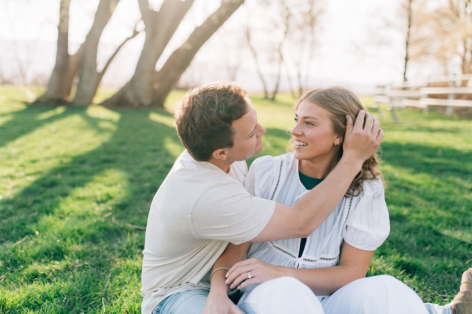 A couple sits together in a grassy park on a sunny day, sharing an affectionate moment under trees.