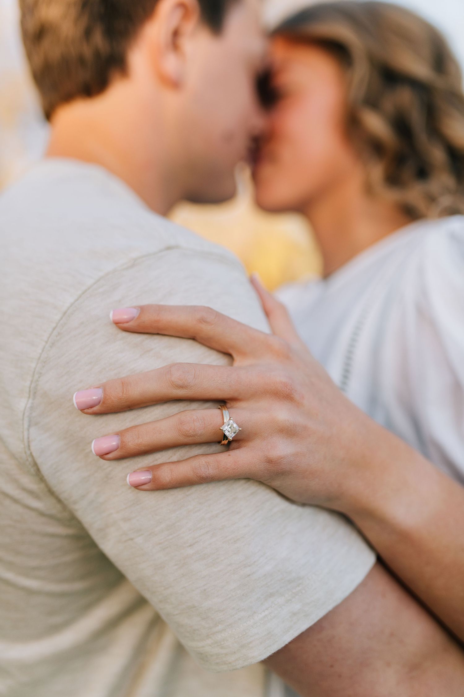 A close-up photo of a couple sharing a kiss outdoors with a diamond engagement ring prominently displayed on her hand.