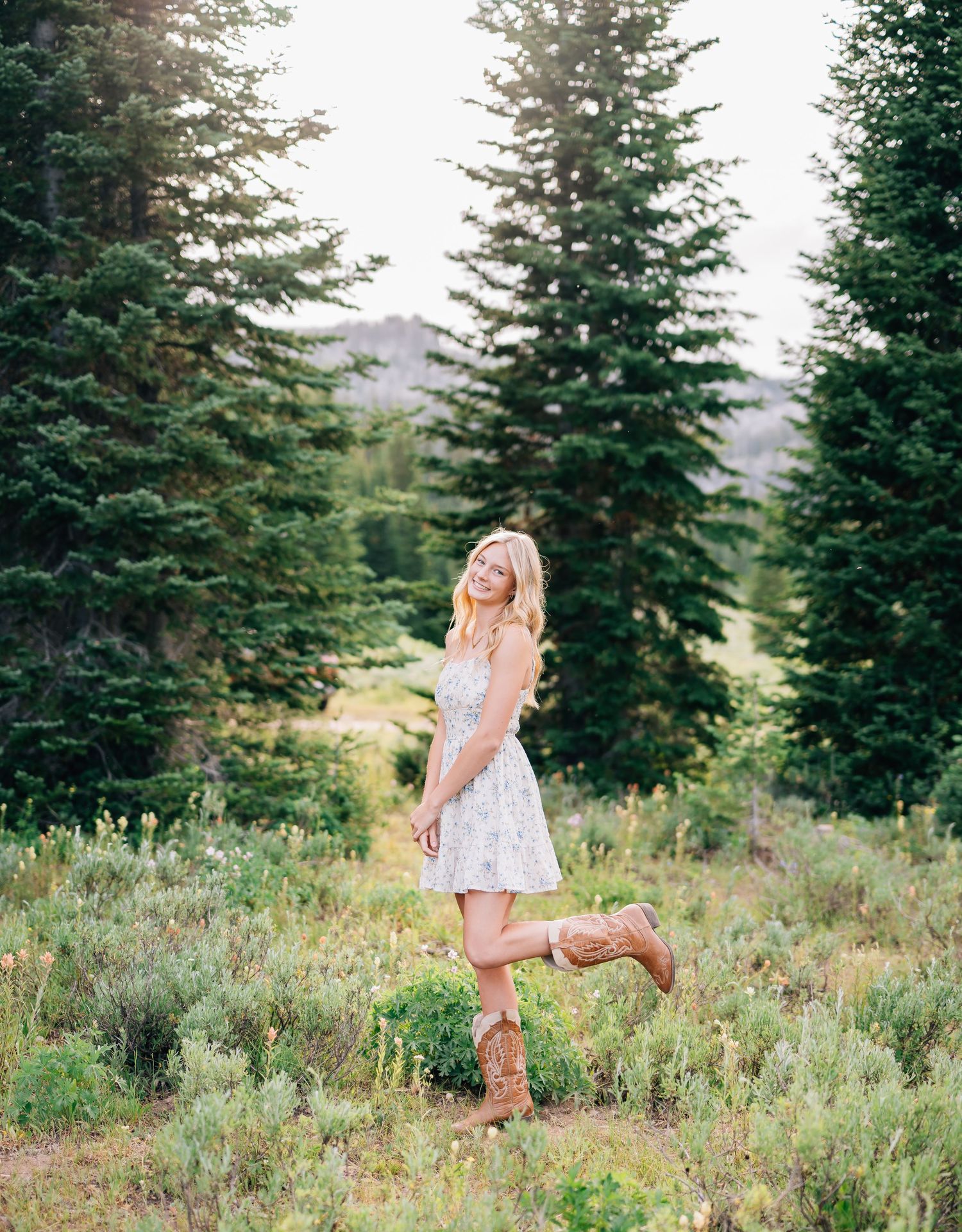 Young woman in floral mini dress and cowboy boots posing in a mountain meadow surrounded by tall pine trees.