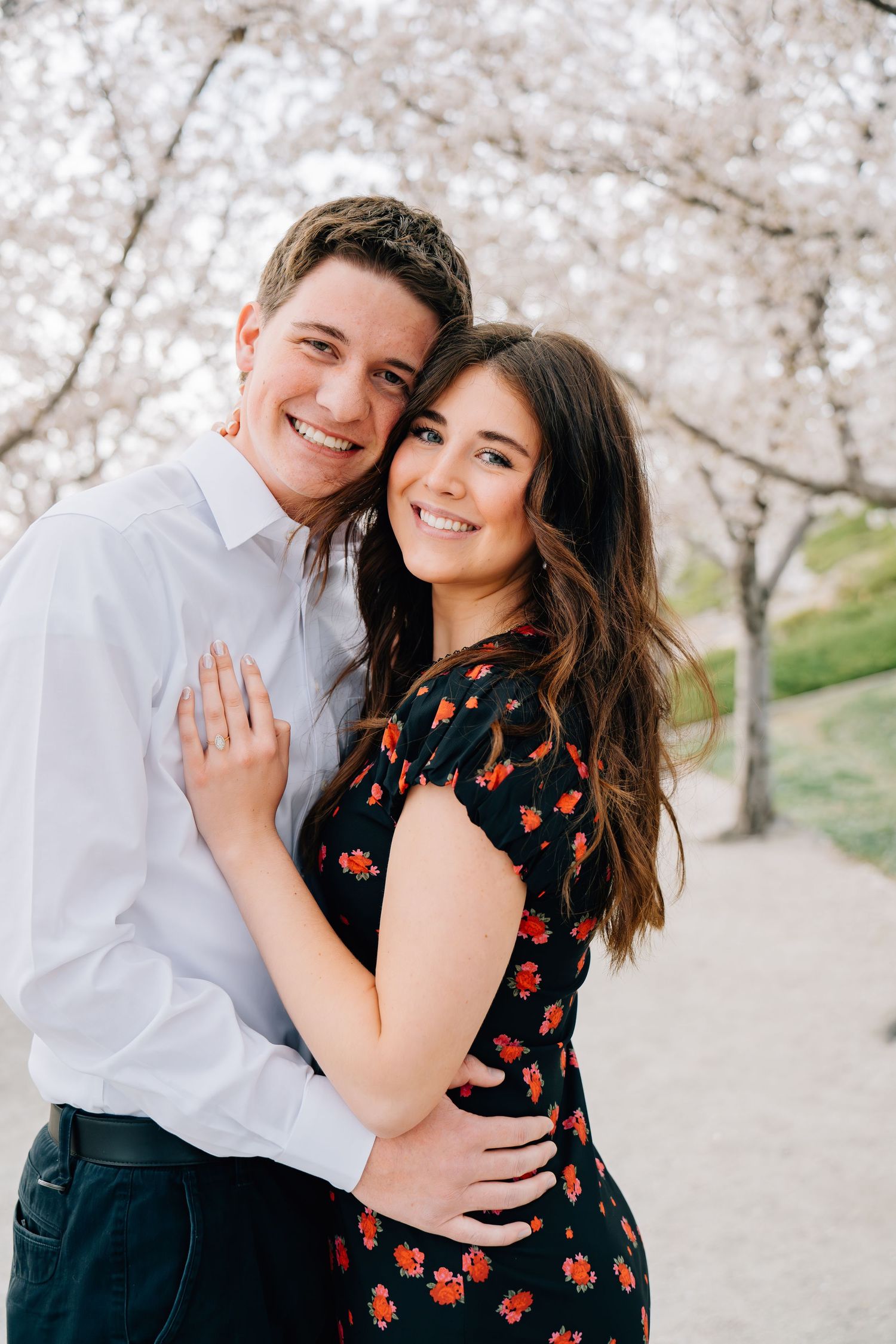 Smiling couple embracing outdoors under blooming cherry blossom trees, woman in floral dress, man in white shirt.