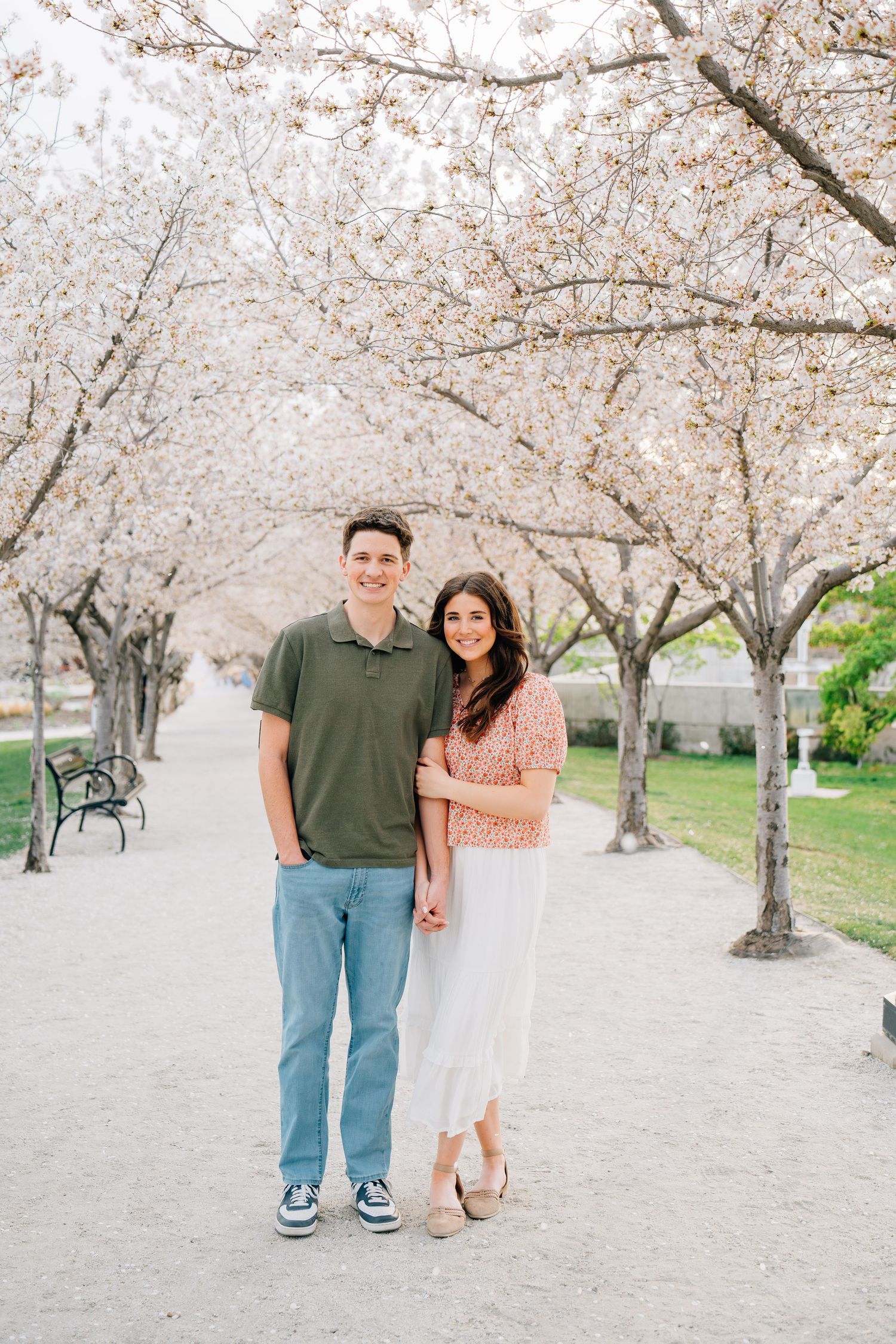 Couple smiling under blooming cherry blossom trees on a park path in spring.