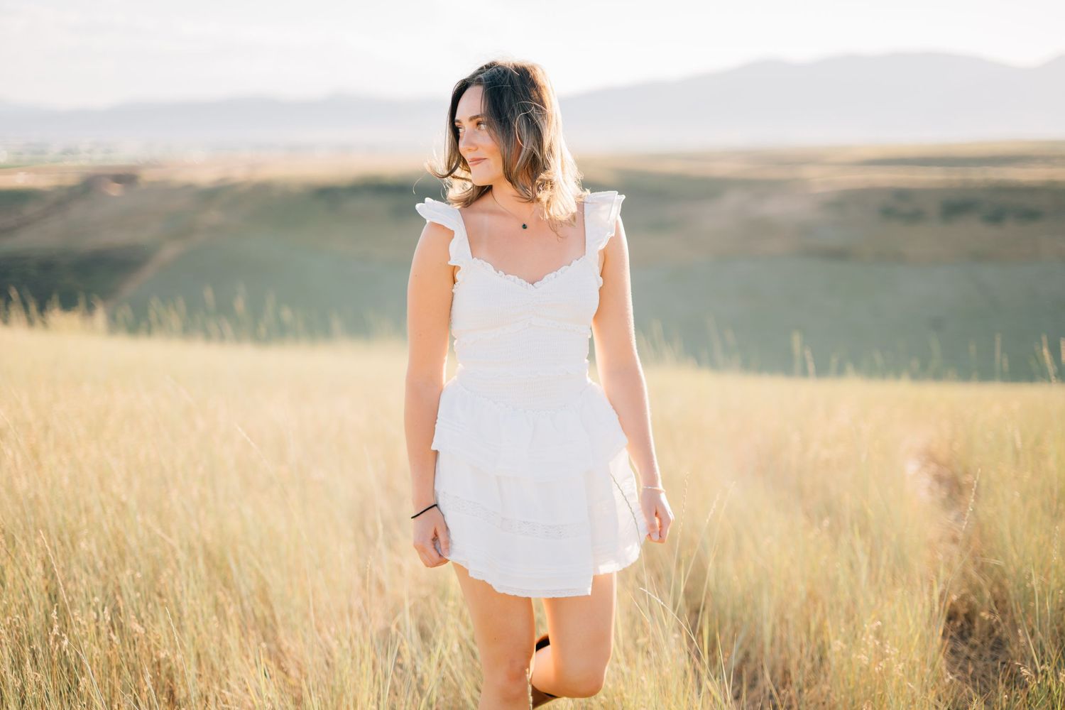 Young woman in white ruffled mini dress standing in golden grass field with mountain backdrop.