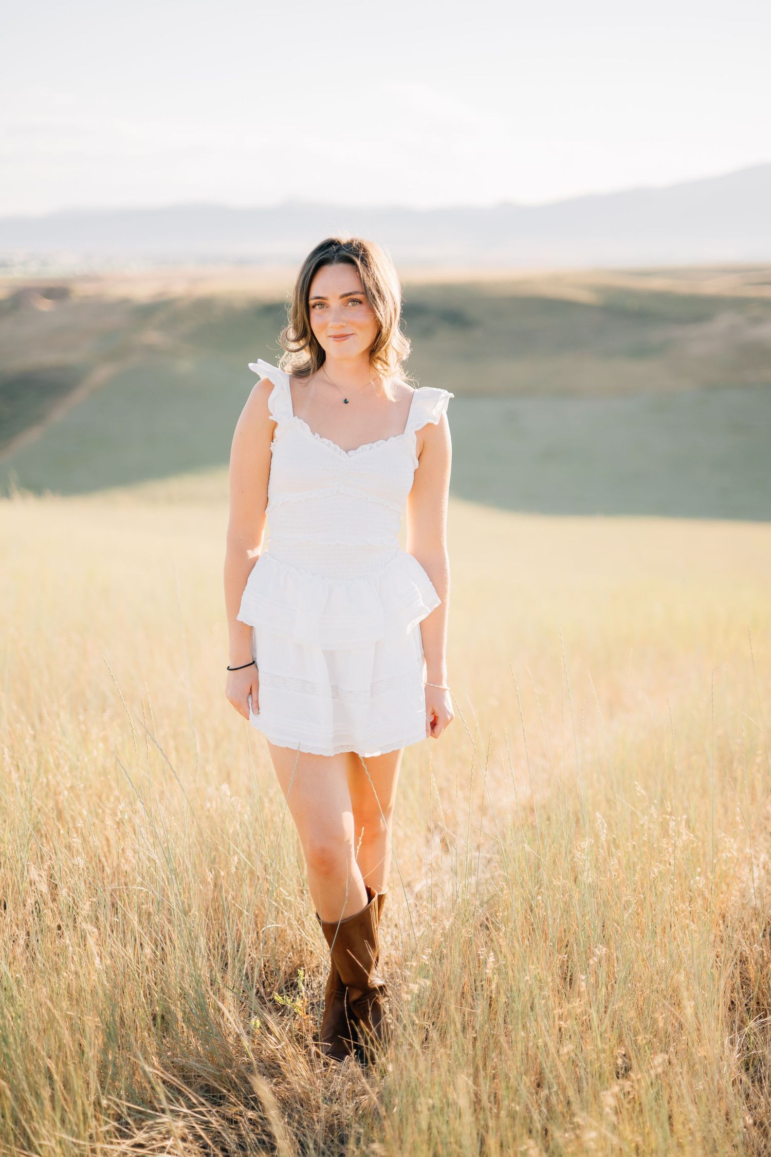Woman in white ruffled mini dress and brown cowboy boots standing in golden field with rolling hills backdrop.