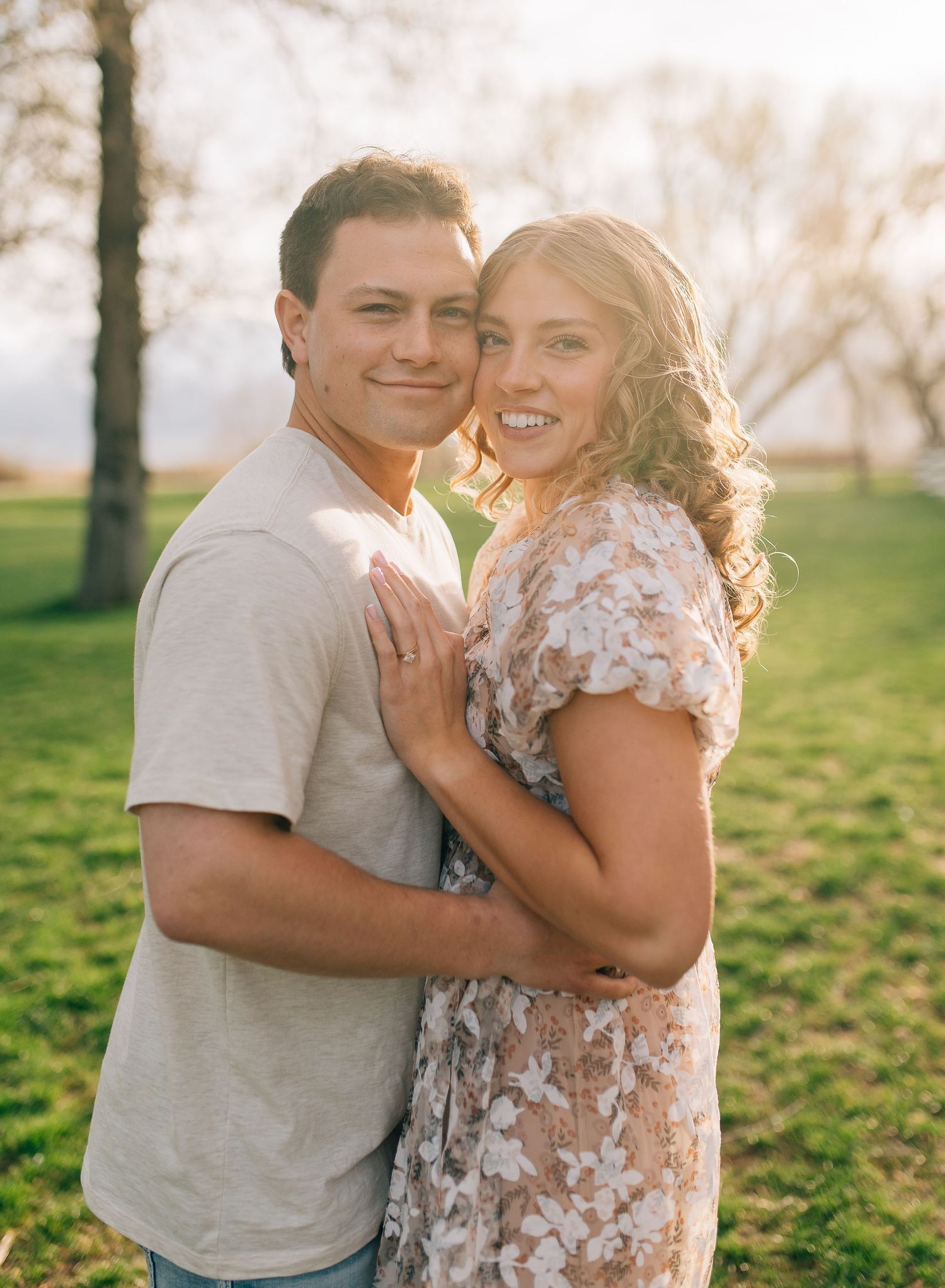Couple embraces on a sunny day in a grassy park during a romantic photoshoot in warm light.