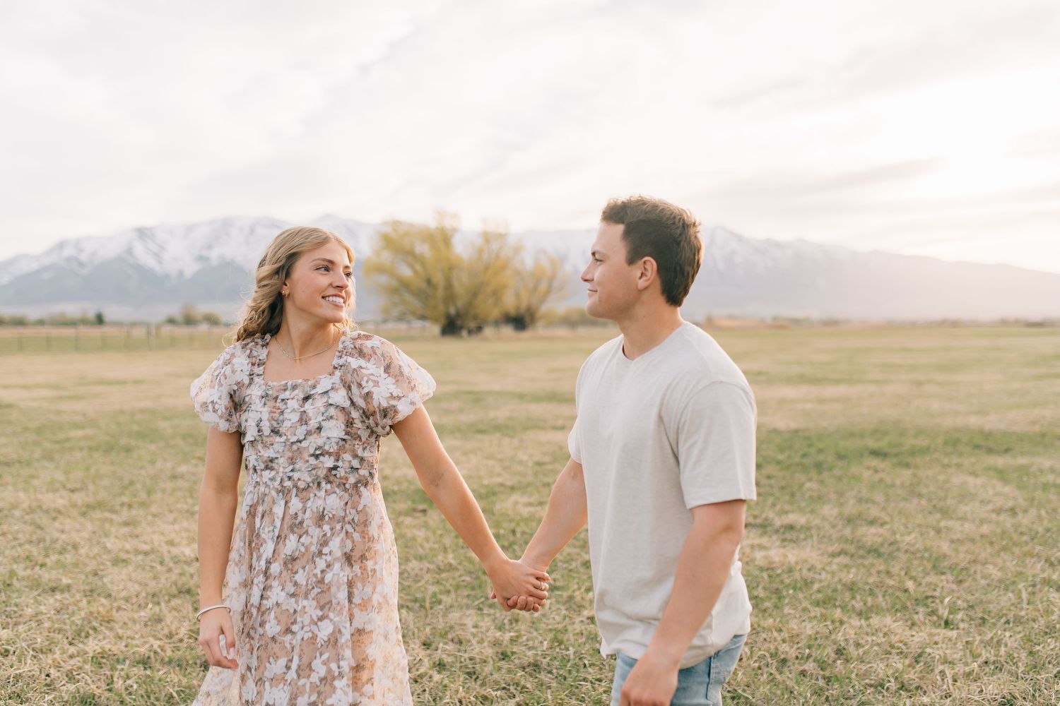 Couple holding hands while enjoying a moment together in an open field with mountain landscape behind them.