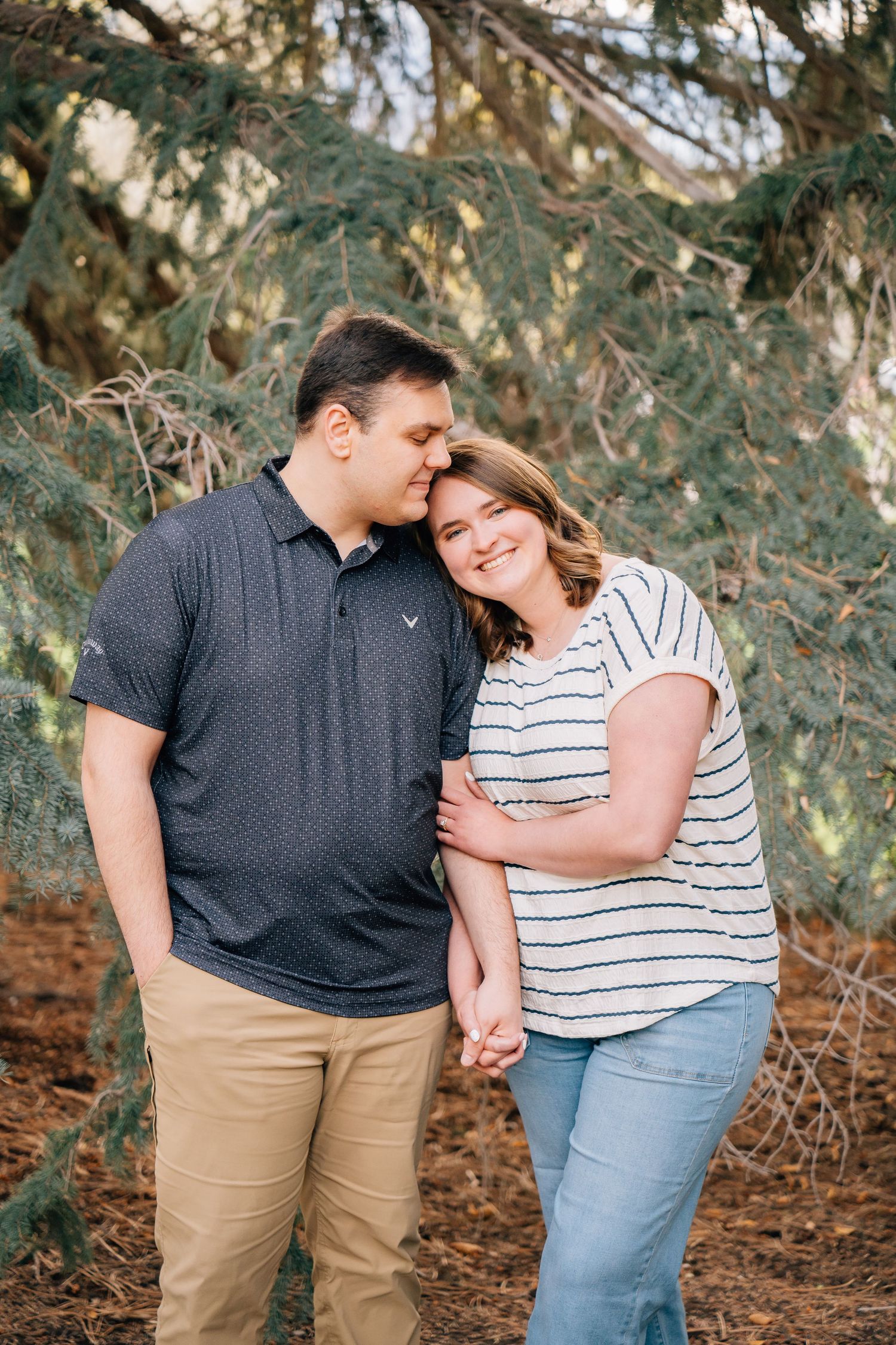 A couple stands under pine trees, holding hands as the man kisses the woman's head in a romantic outdoor portrait.