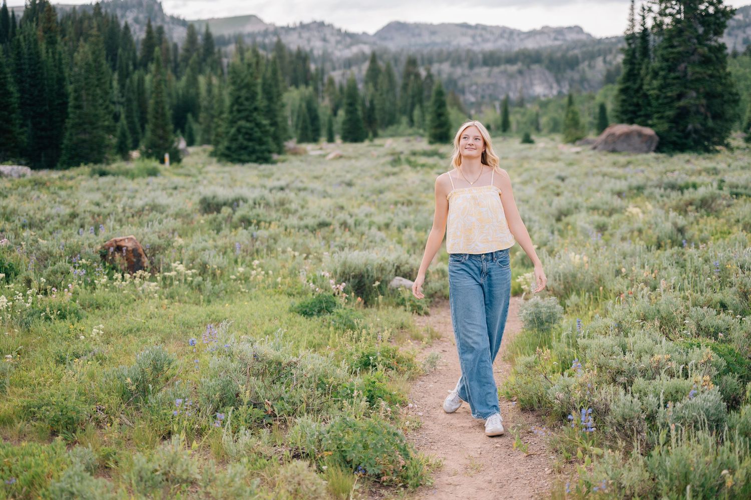 Woman walking on a mountain meadow trail surrounded by wildflowers and pine trees with rocky peaks in the background.