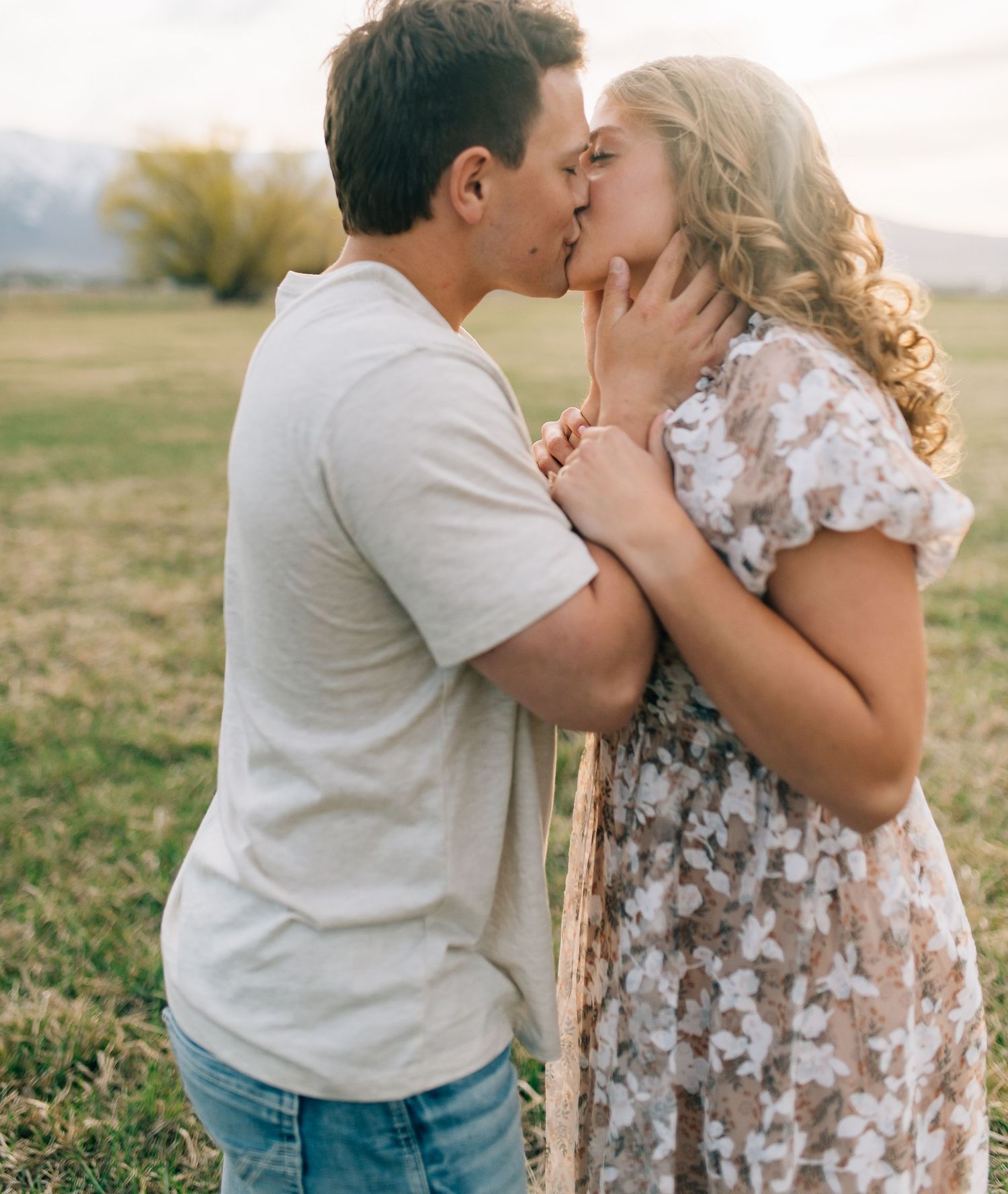 A couple shares a tender kiss in a floral dress and casual attire while standing in a sunlit grassy field.