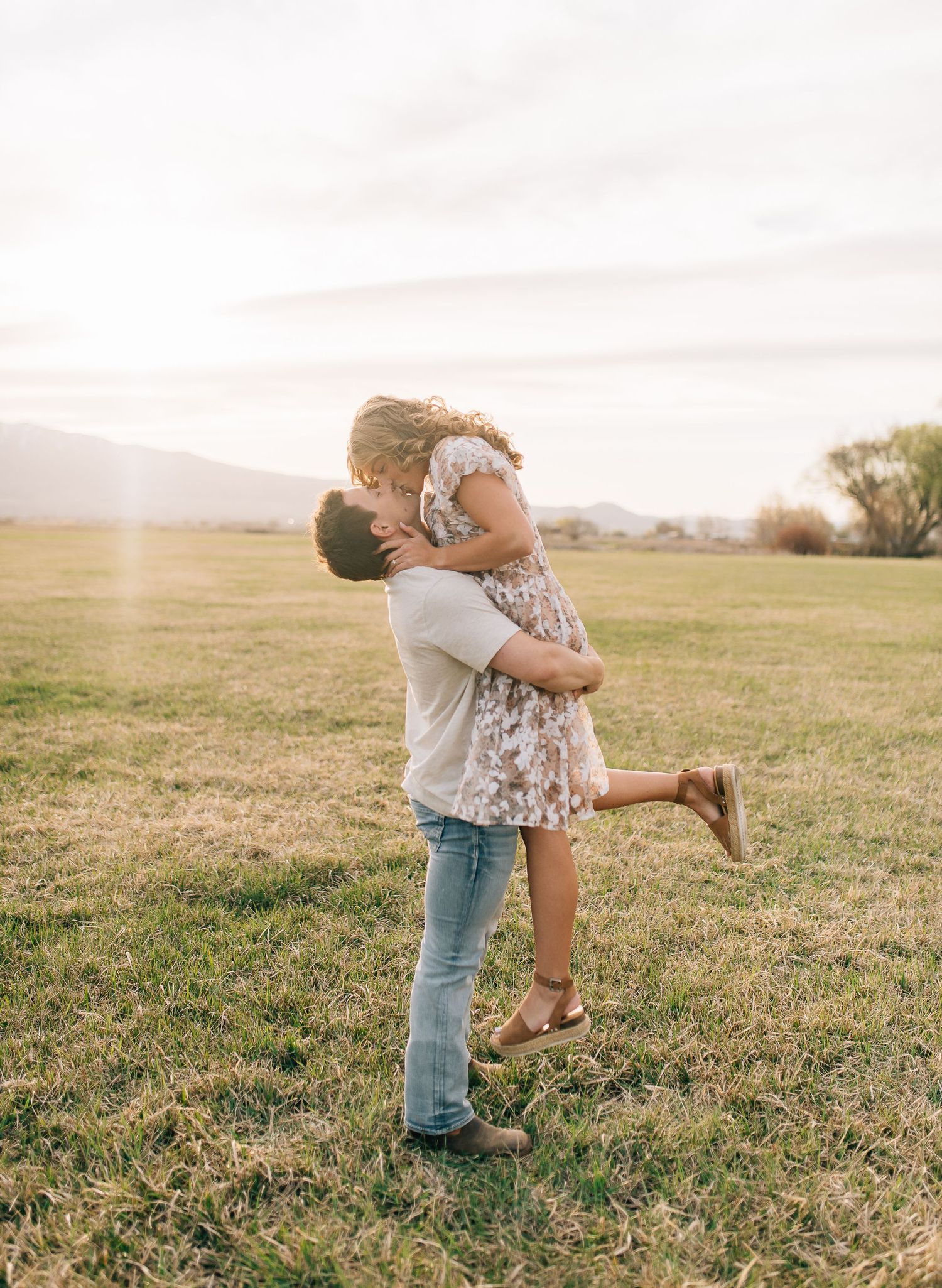A couple embrace in a loving hug on a grassy field at sunset with mountains in the distance.