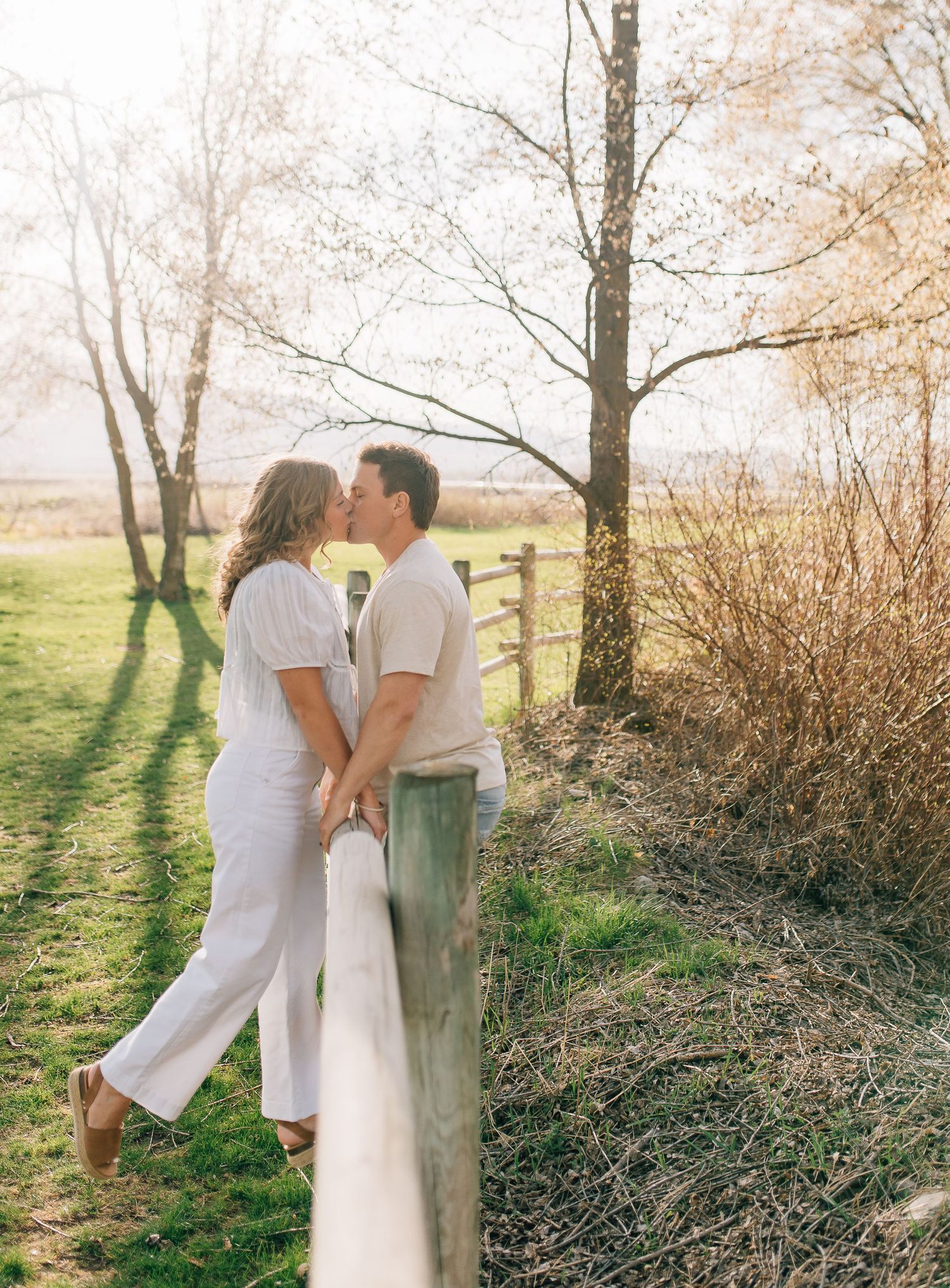 Couple in white outfits sharing a romantic moment while leaning against a wooden fence in sunlit countryside setting.