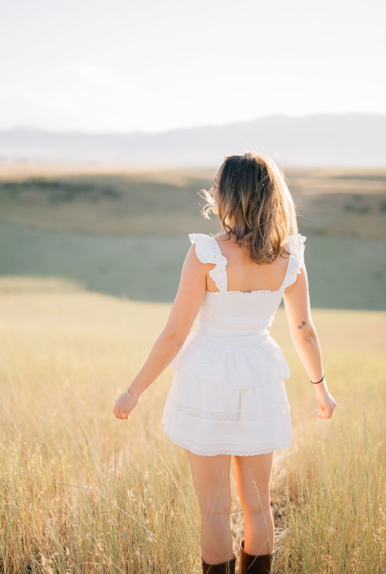 Woman in white ruffle mini dress and brown boots standing in golden field with mountain backdrop.