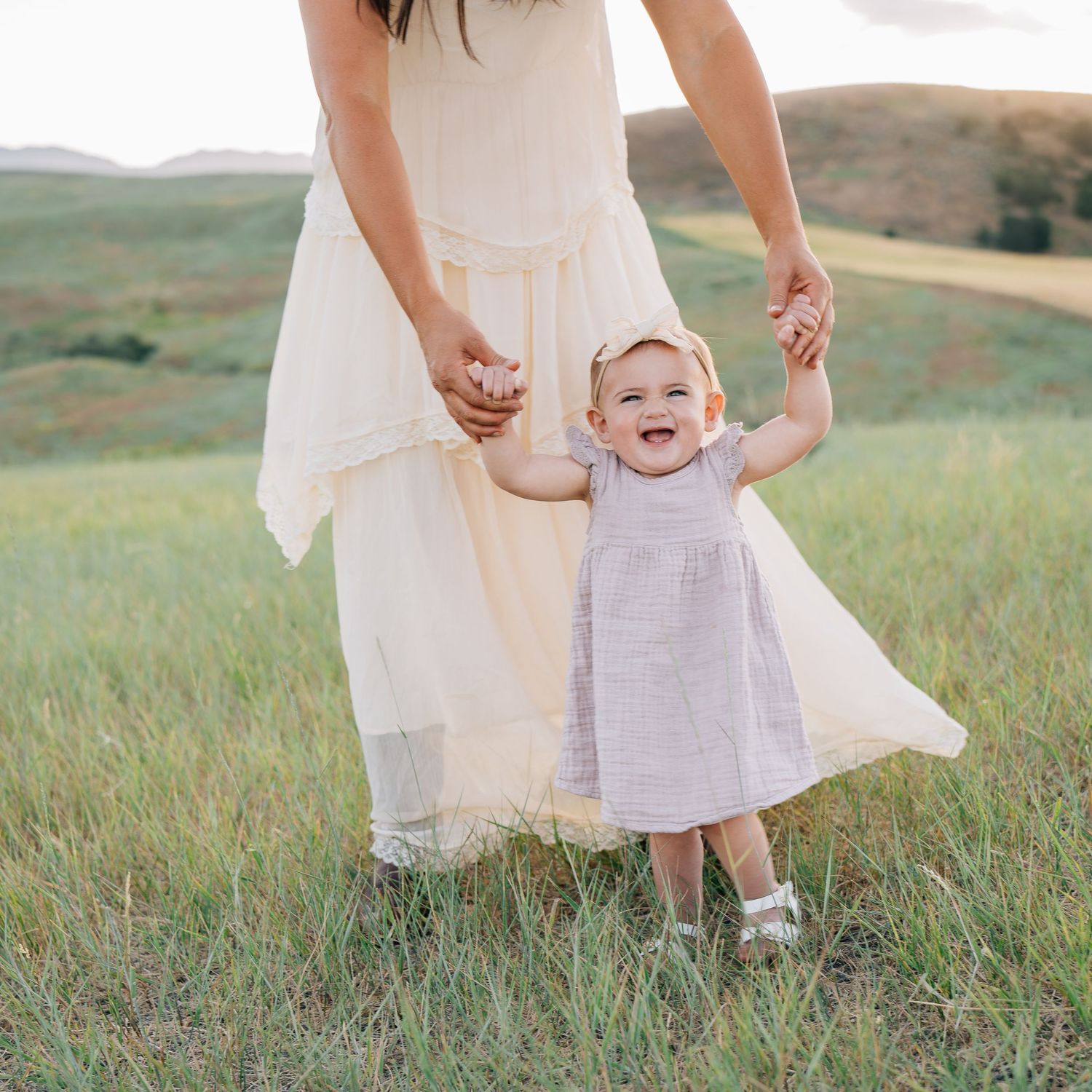 A smiling baby in a lavender dress holds her mother's hands while standing in a green meadow with rolling hills.