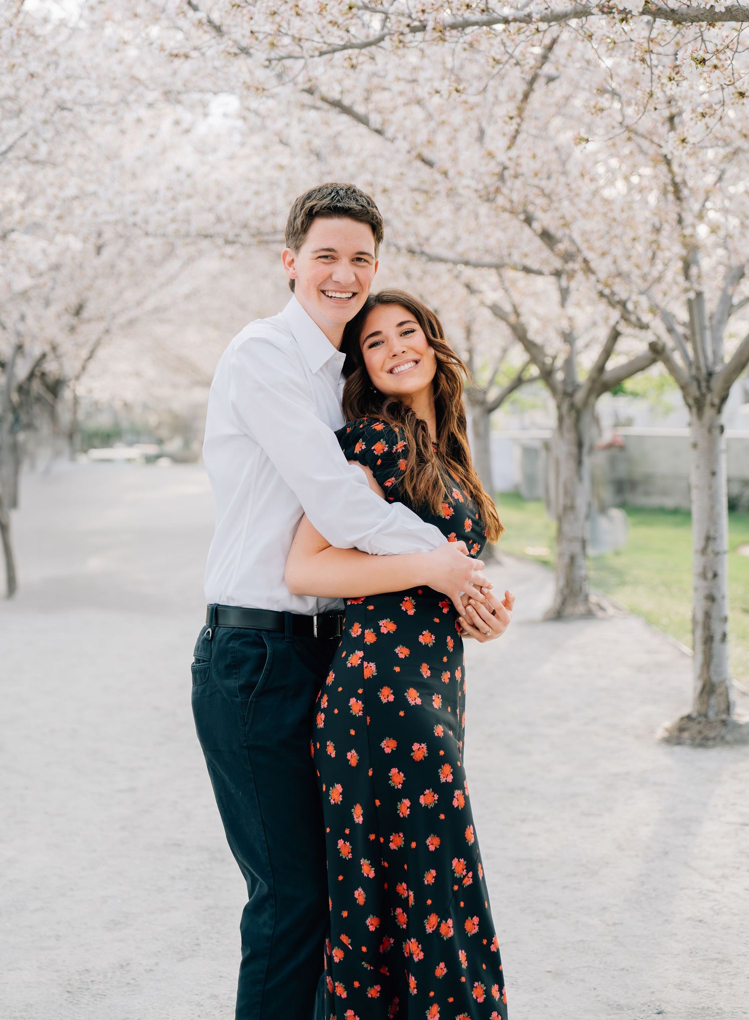 Couple embracing under blooming cherry blossom trees, woman in floral dress, man in white shirt and dark pants.