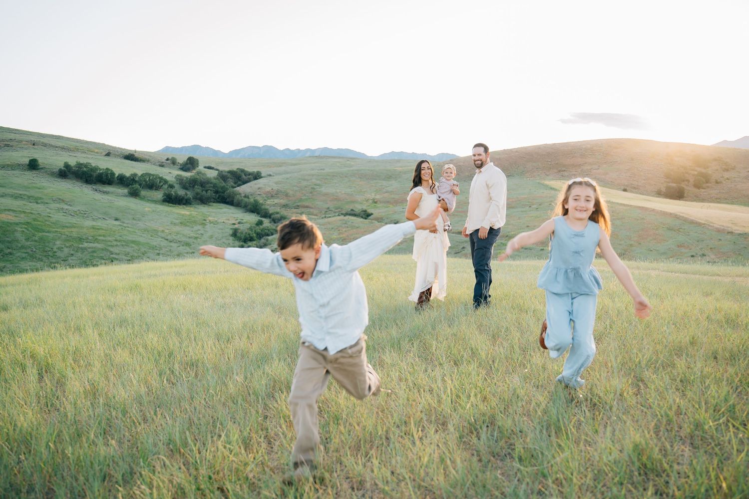 Family of four enjoying a sunny day in green hills, two children running with arms out while parents hold a baby.