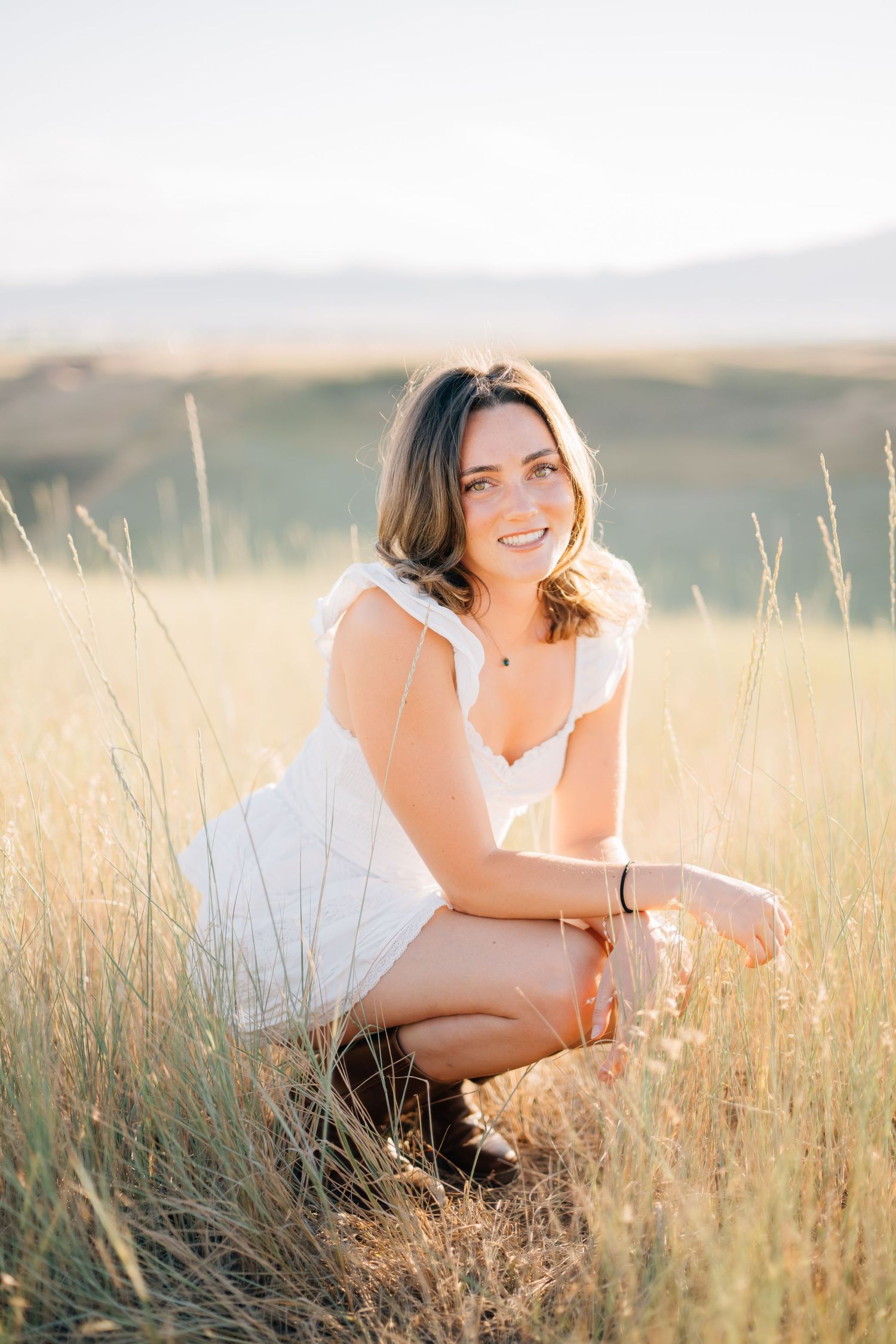 Young woman in white dress crouching in golden grass field, smiling warmly with mountains in background.