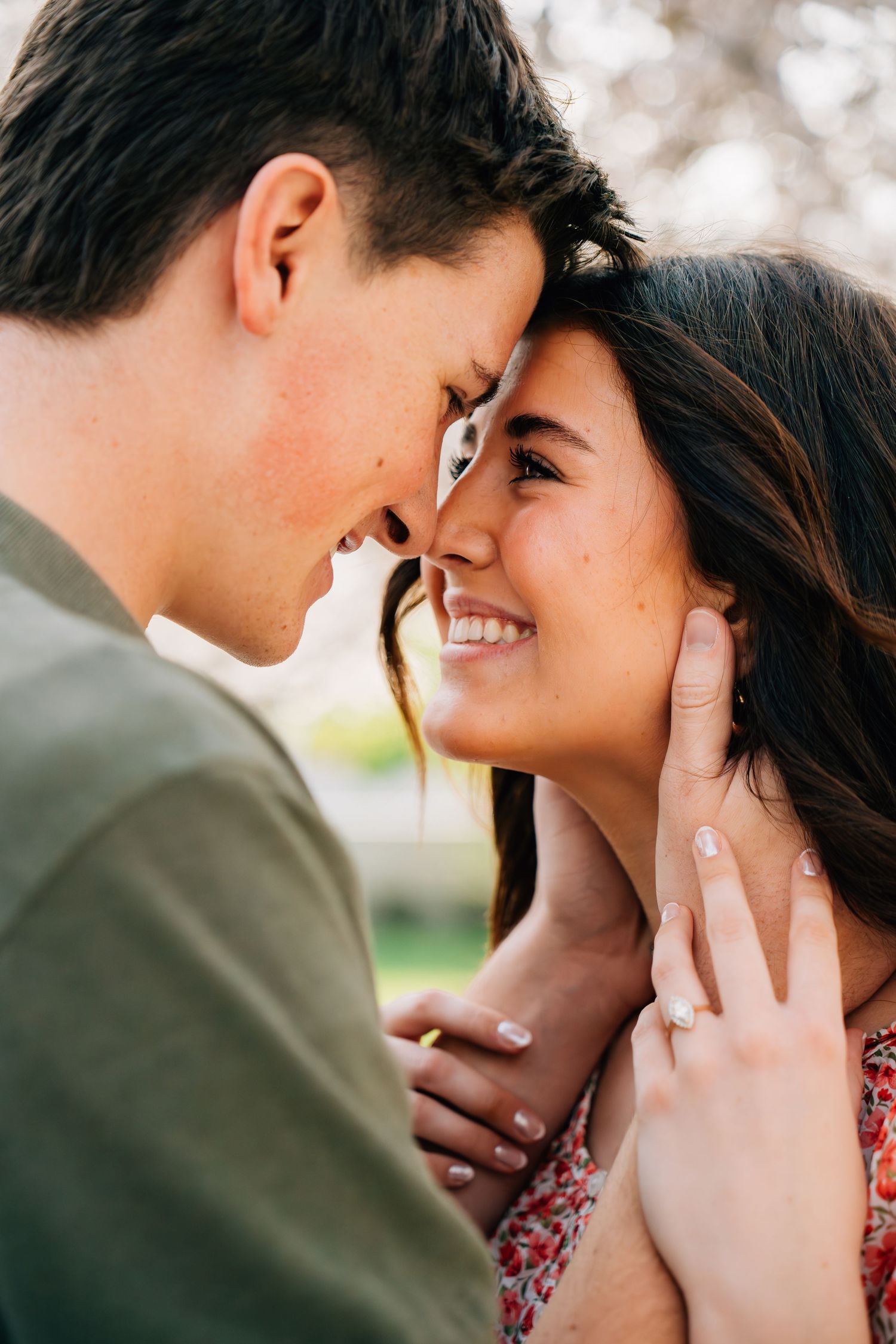 A smiling couple in a romantic forehead-to-forehead pose outdoors, with an engagement ring visible on the woman's hand.