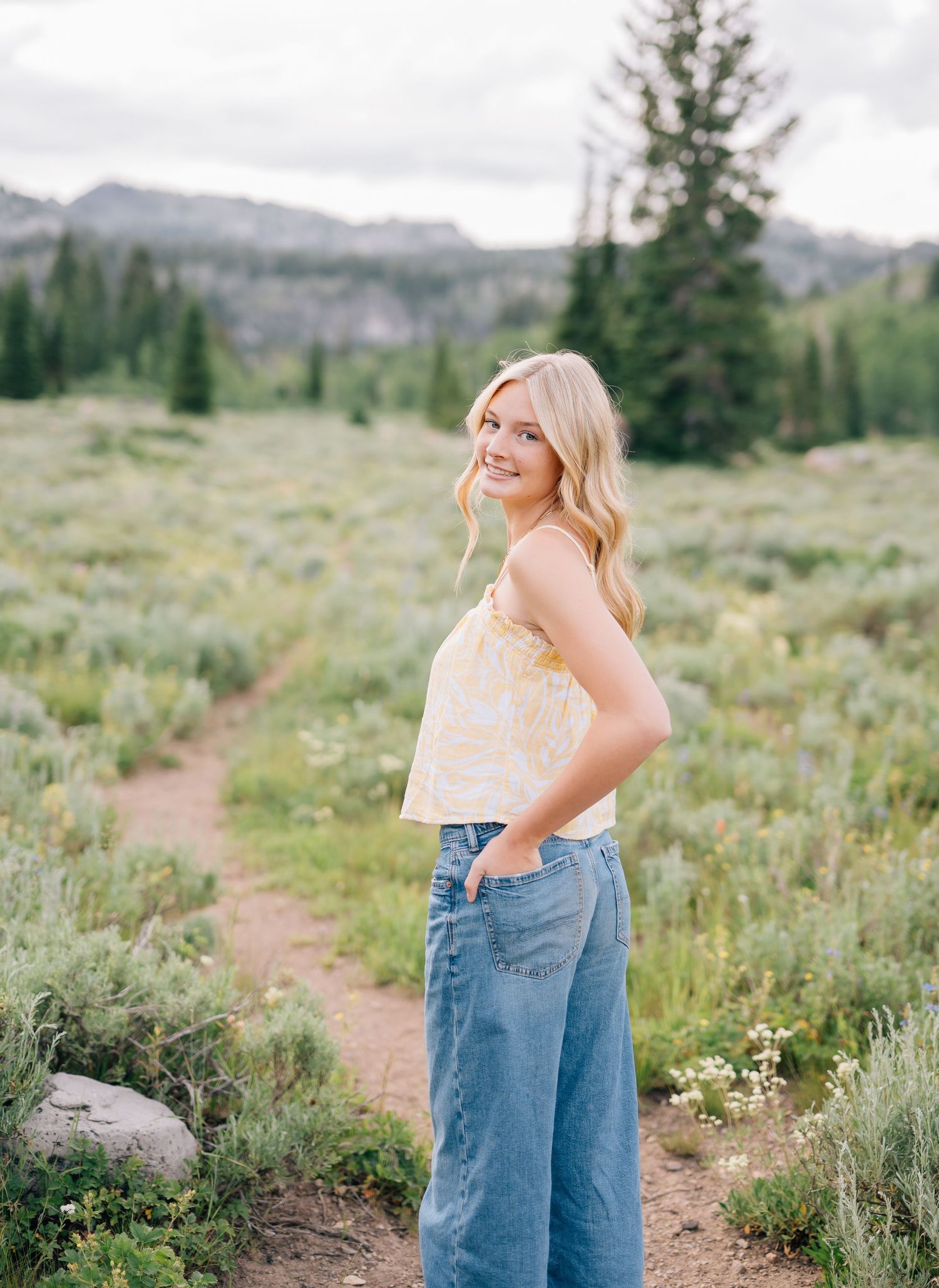 Smiling blonde woman in yellow floral top and wide-leg jeans poses on a mountain meadow trail surrounded by greenery.