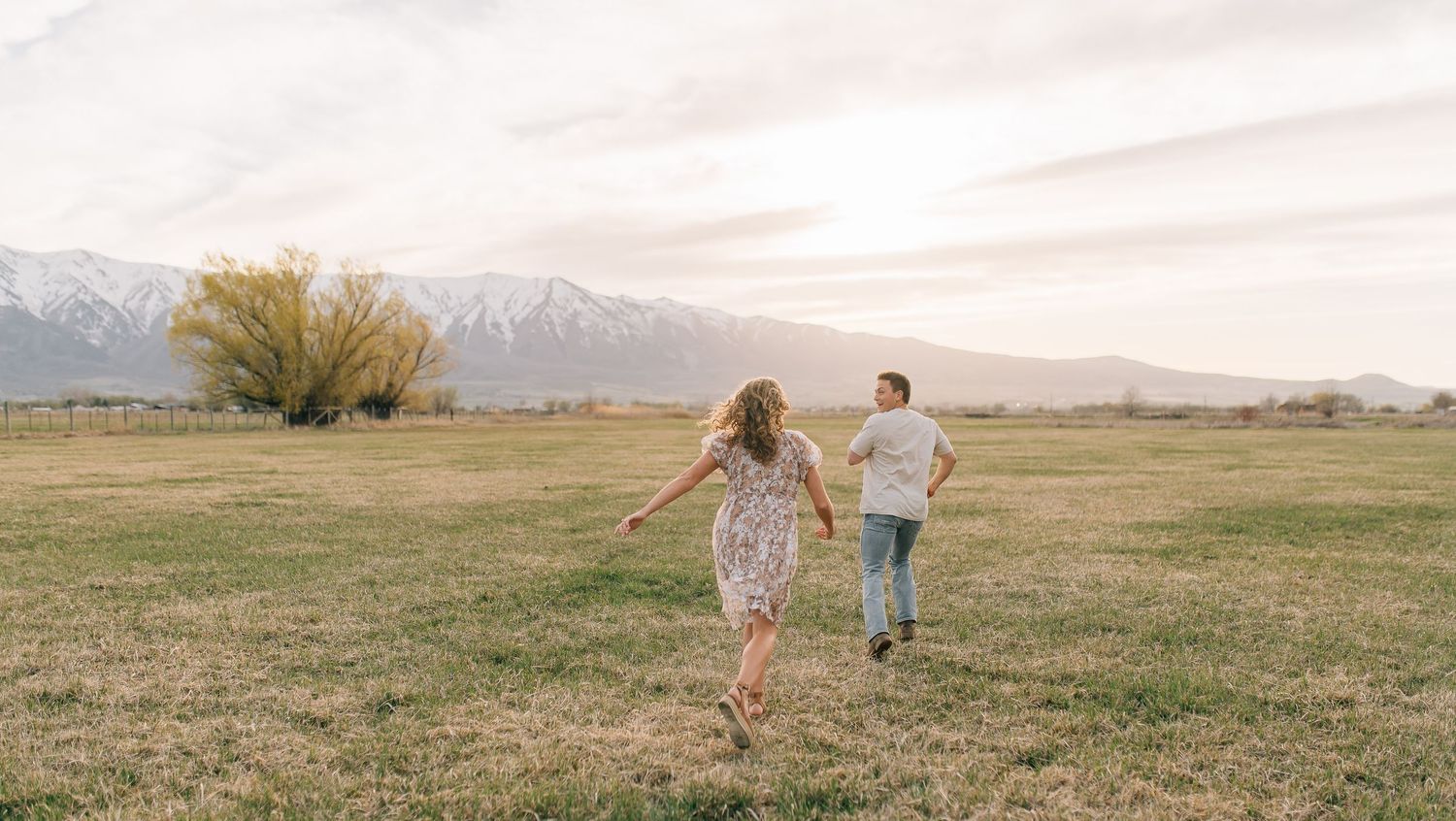 Couple running playfully in a grassy field with snow-capped mountains and autumn trees in the background.