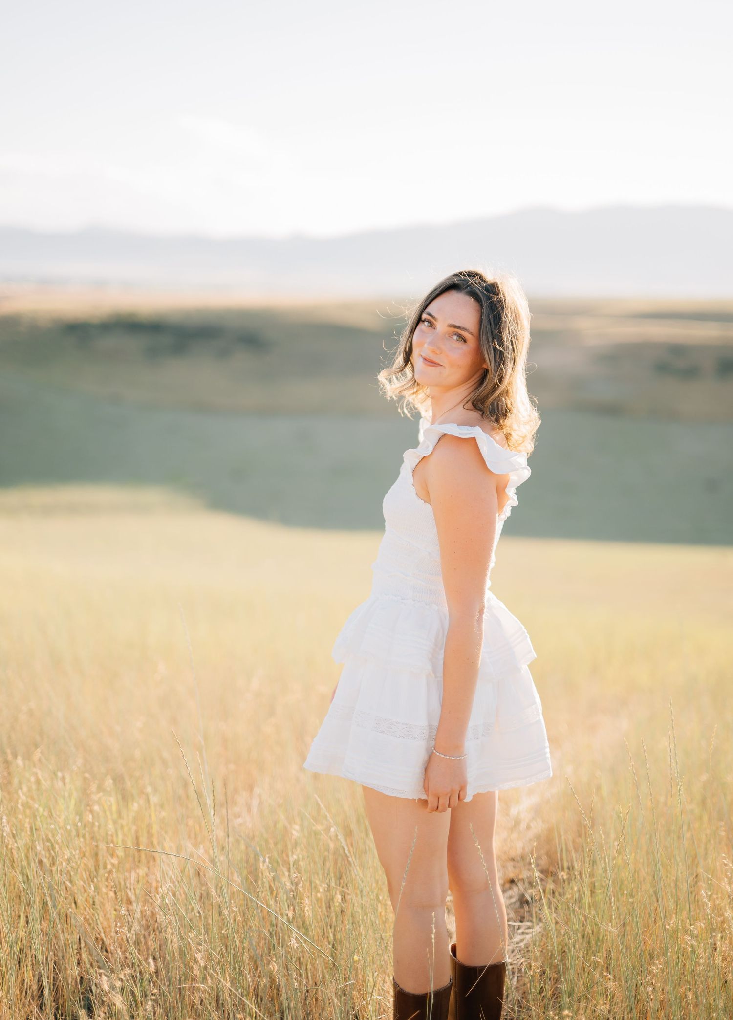 Young woman in white dress and brown boots standing in golden field with mountain backdrop in warm sunlight.