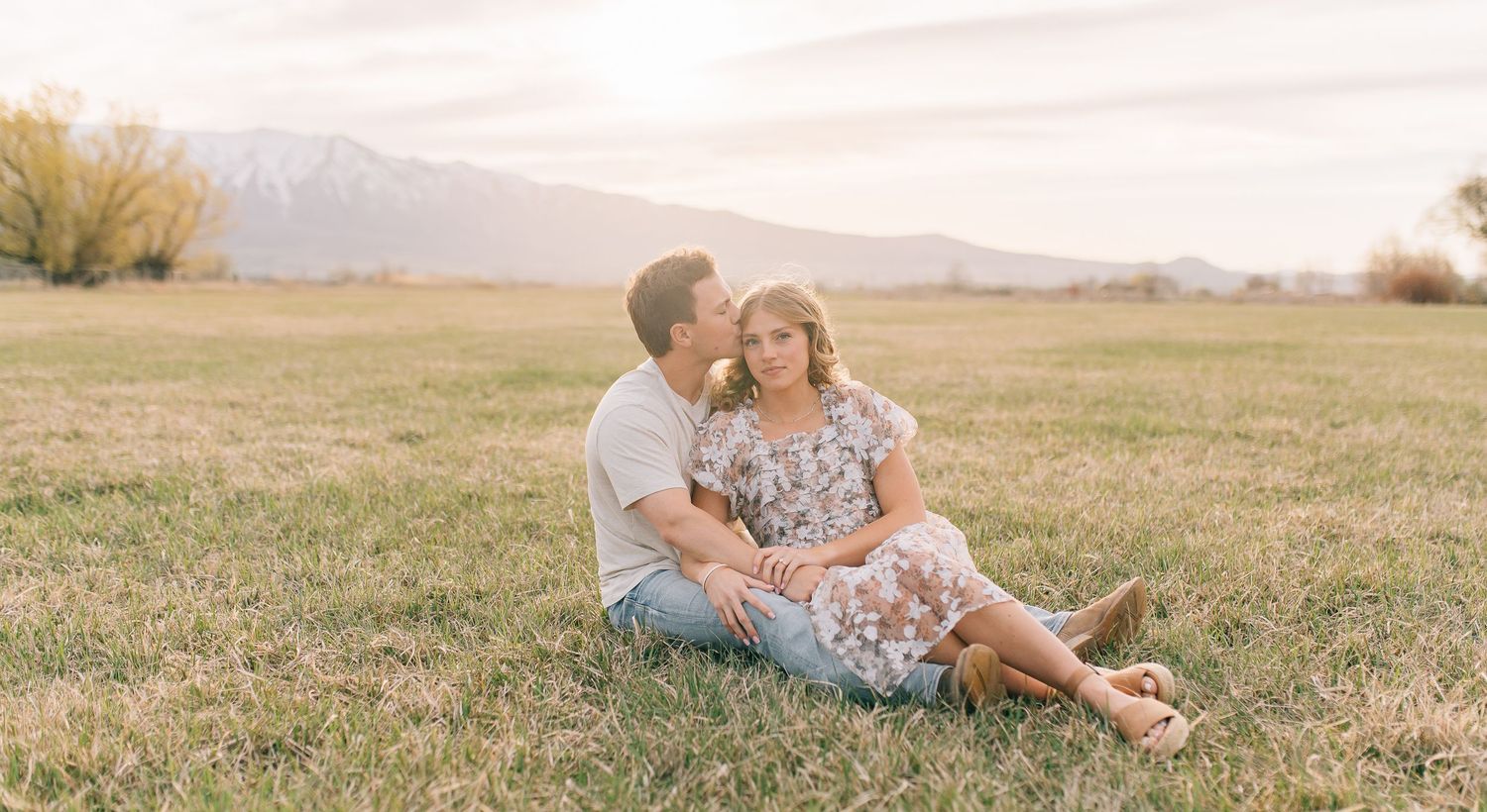 A couple sits together in a sunny meadow with mountains visible in the background during golden hour.