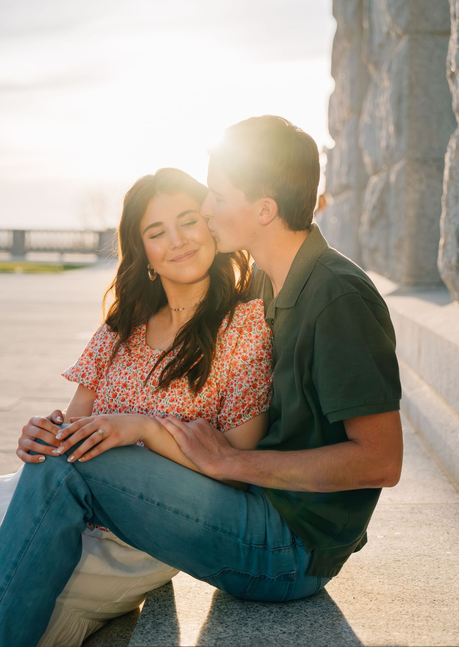 A couple shares a romantic moment at sunset, man kissing woman on cheek while sitting outdoors.